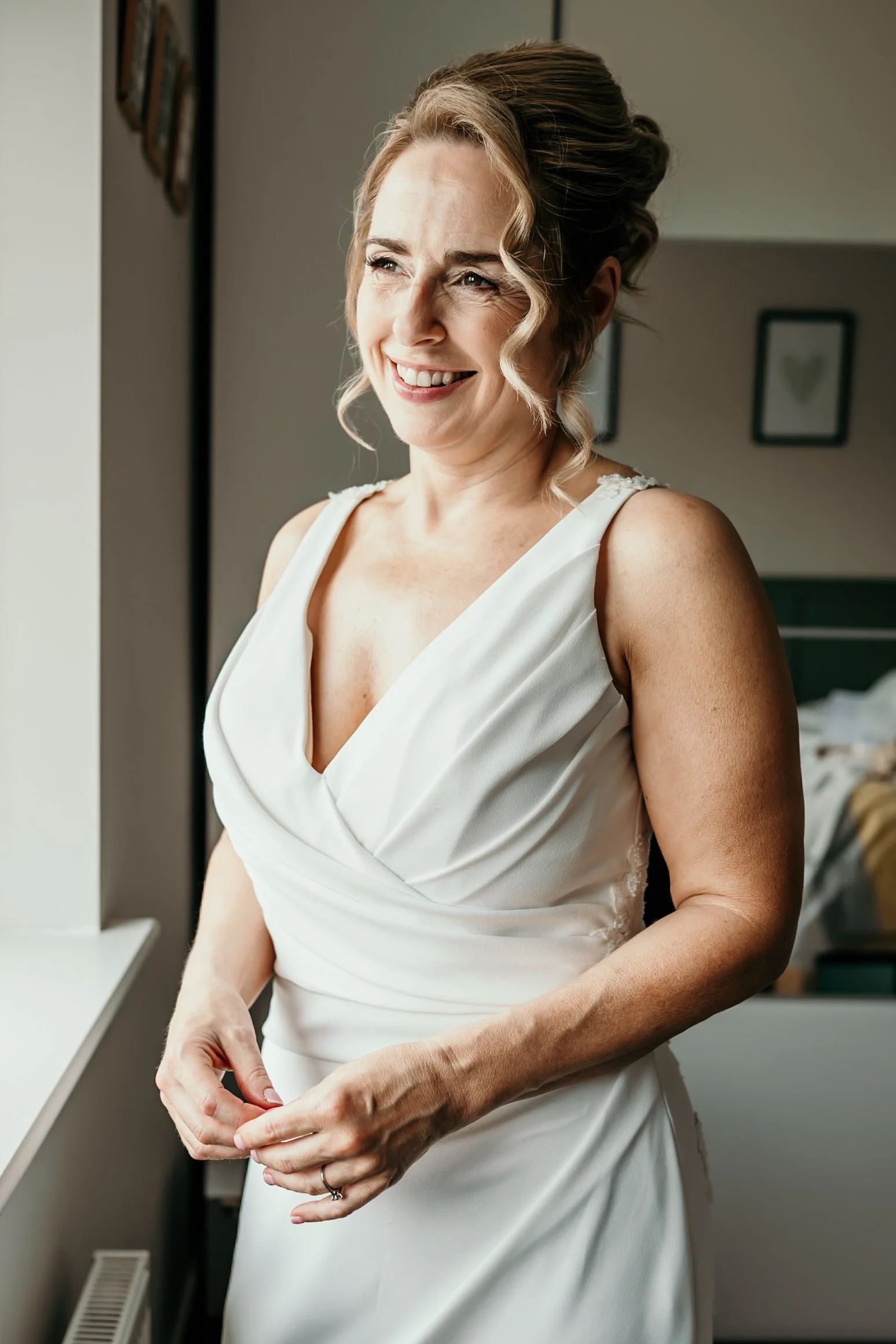 A bride stands in her wedding dress looking out the window and smiling