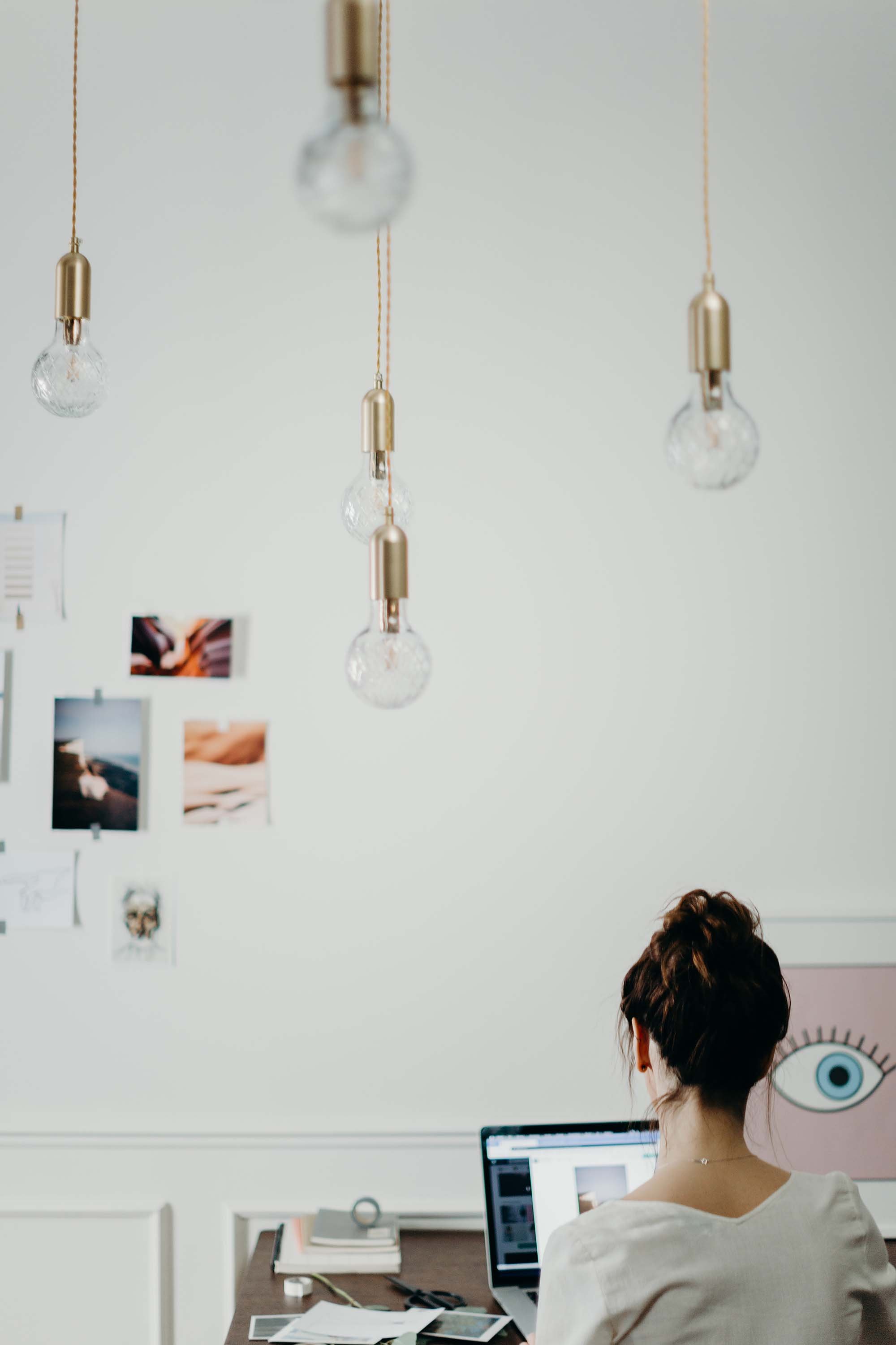 Mujer trabajando en una oficina con luz colgante y pared decorada con fotos y dibujos.