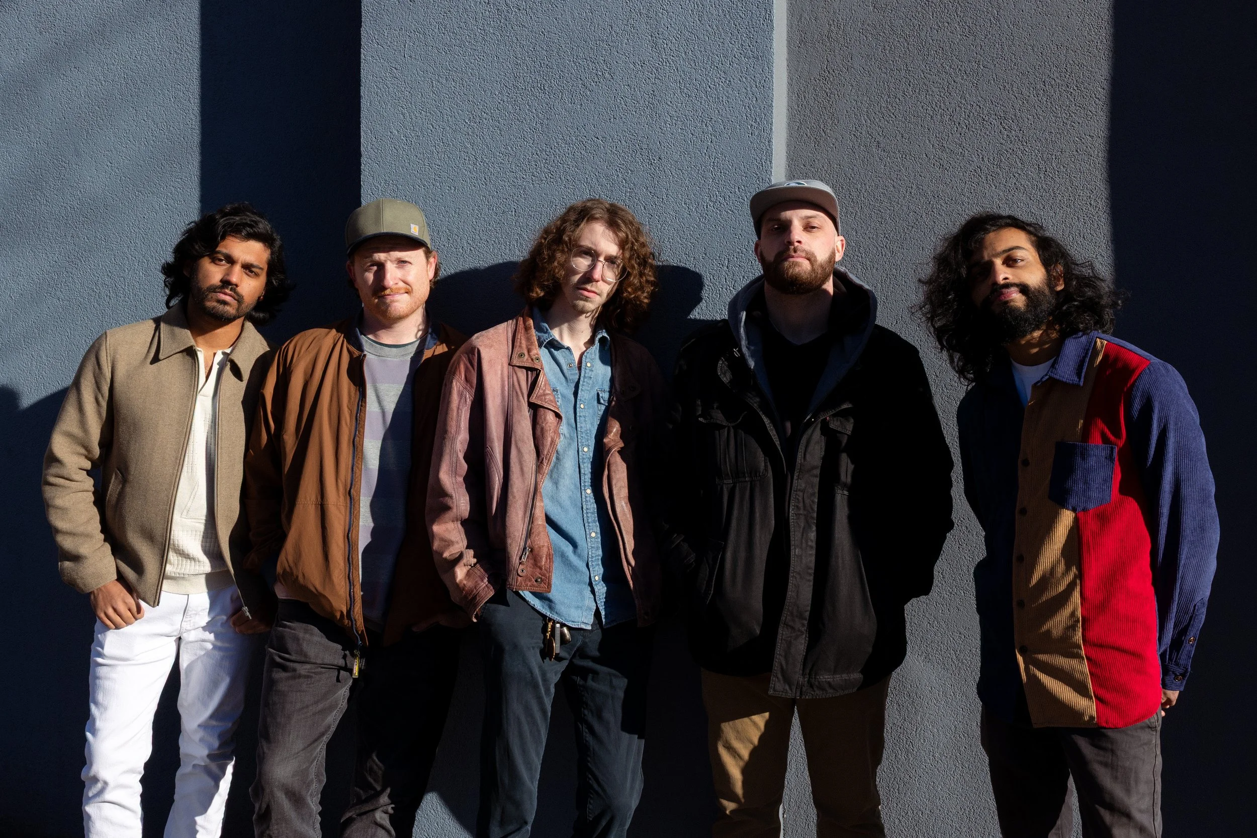 Group of five men standing outdoors against a gray wall, with sunlight casting shadows on the wall.