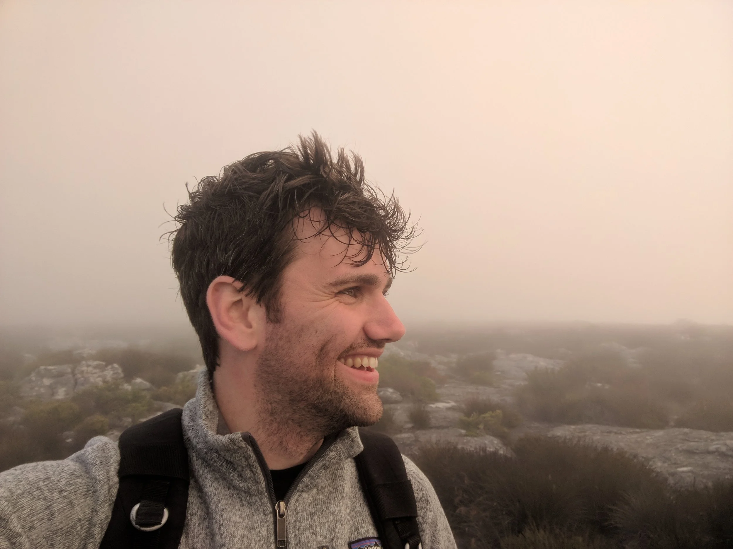 A man smiling and looking to the right, outdoors in a foggy landscape with rocks and sparse vegetation.