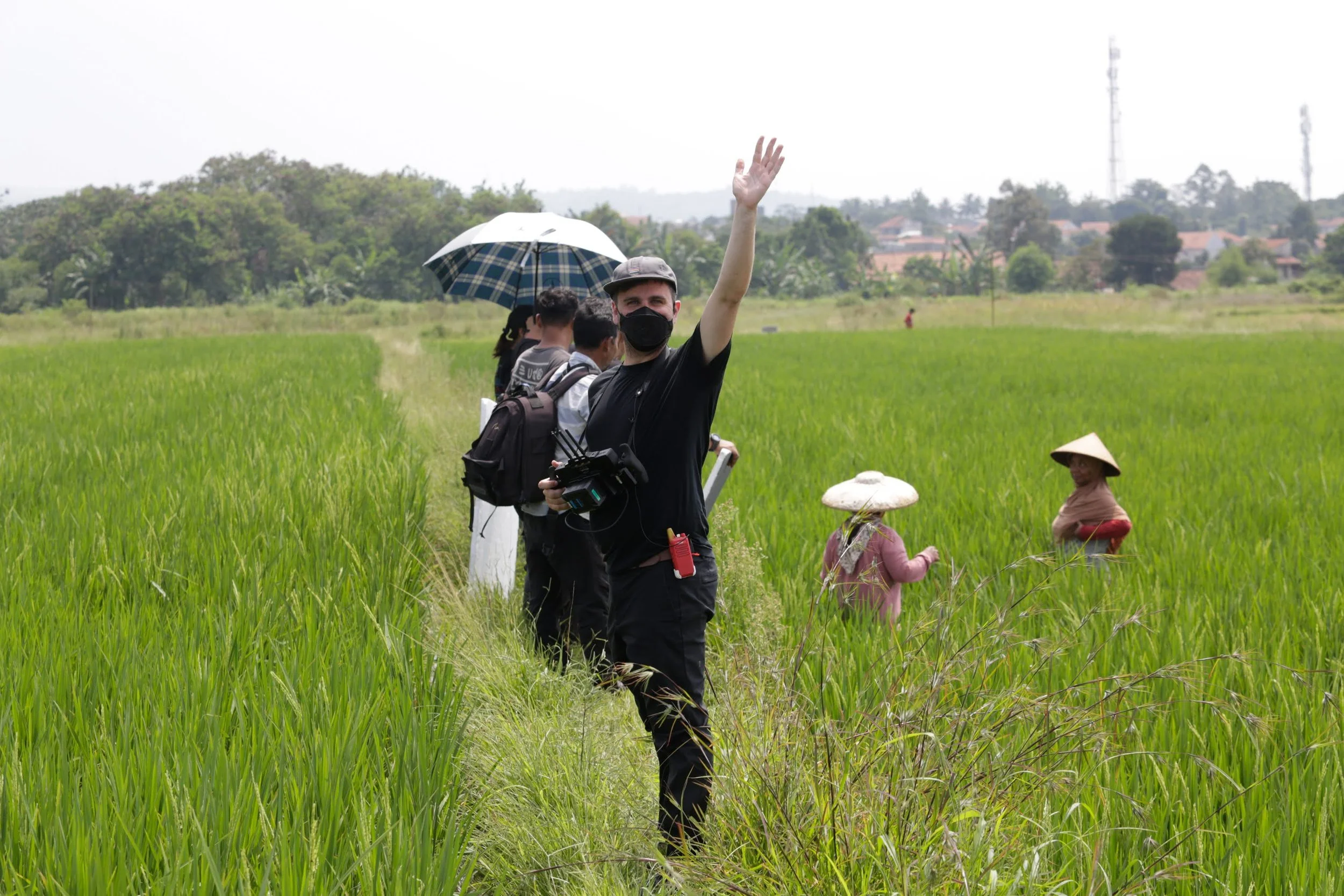 A group of people, including a man with a camera, walking through a green rice field. The man is waving at the camera. Some individuals are using umbrellas and wearing traditional conical hats, likely farmers.