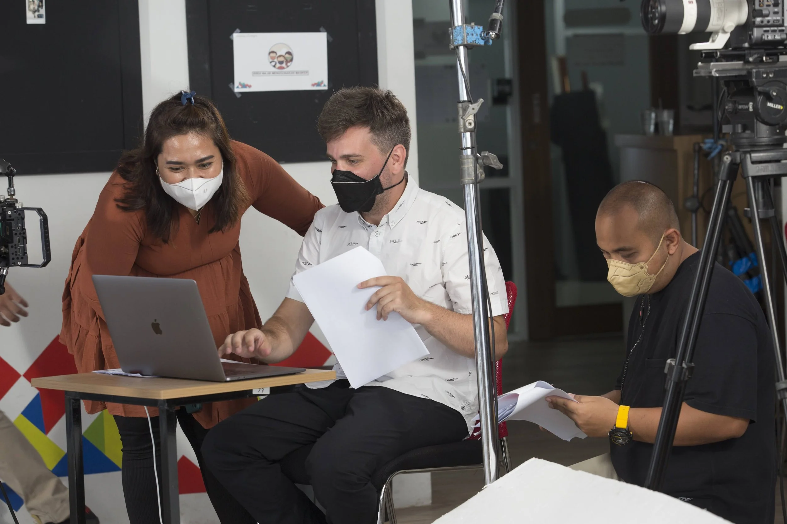 People working on a film or video production set, wearing face masks, with equipment such as cameras and a laptop visible.