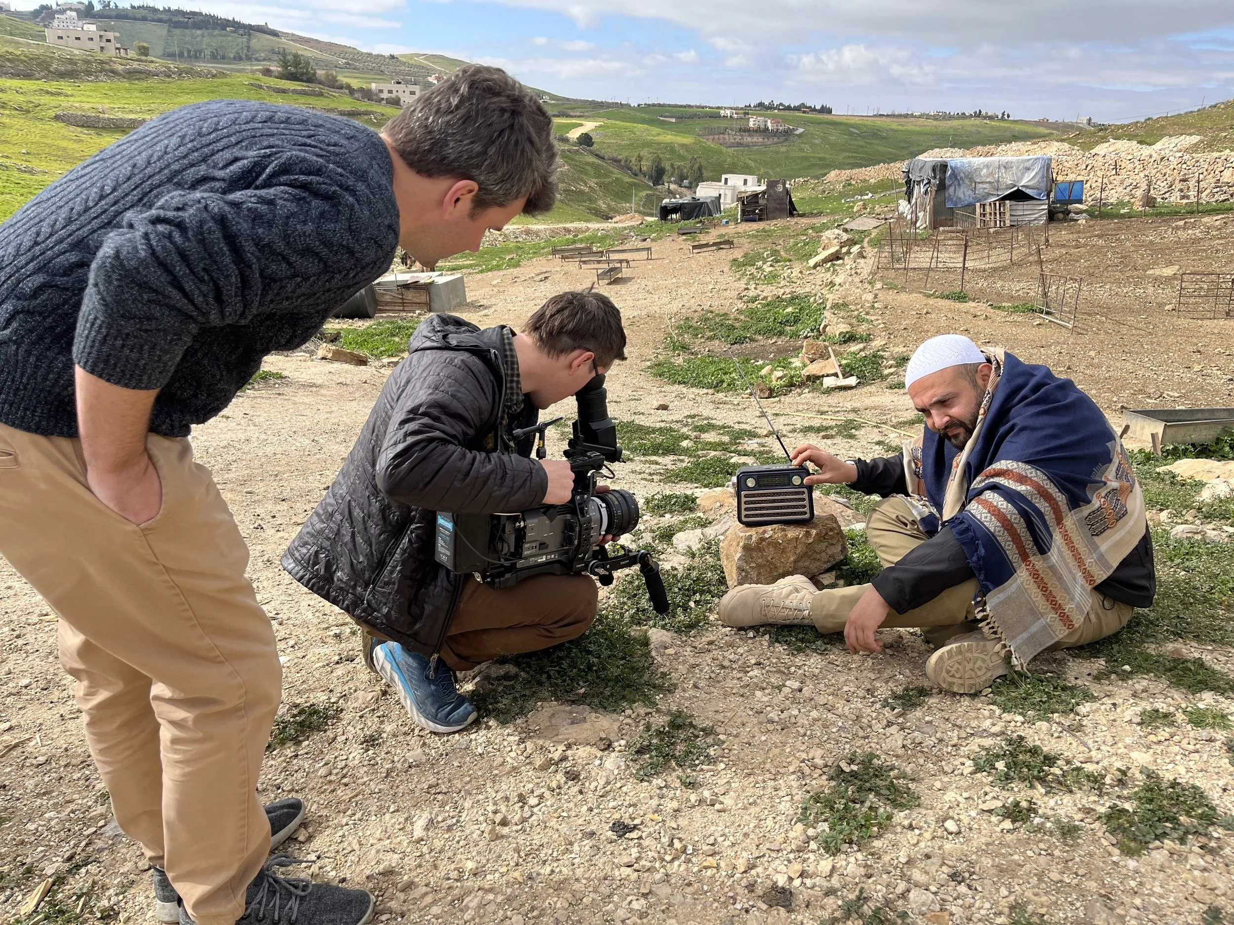 Three men in a rural outdoor setting, one of whom is sitting on the ground with a portable radio, while the other two are standing, one operating a camera, possibly filming or taking photos.
