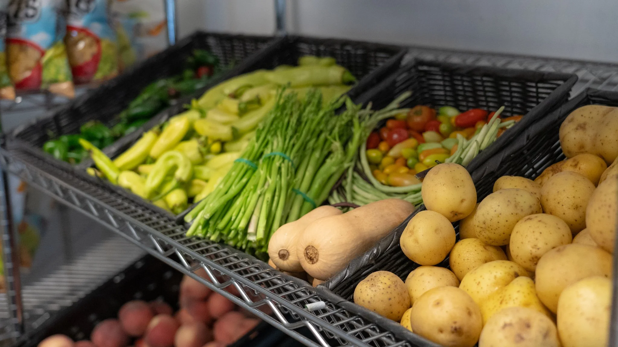 Food Markets and Retail: Black wicker baskets on a metal rack containing peppers, asparagus, tomatoes, squash, and potatoes