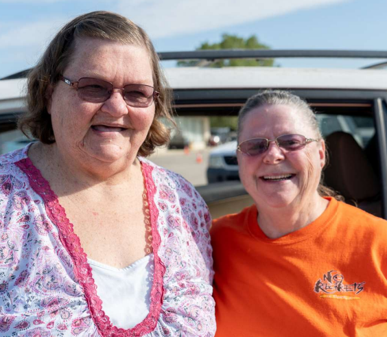 Two smiling people in glasses standing in front of vehicles at mobile pantry distribution