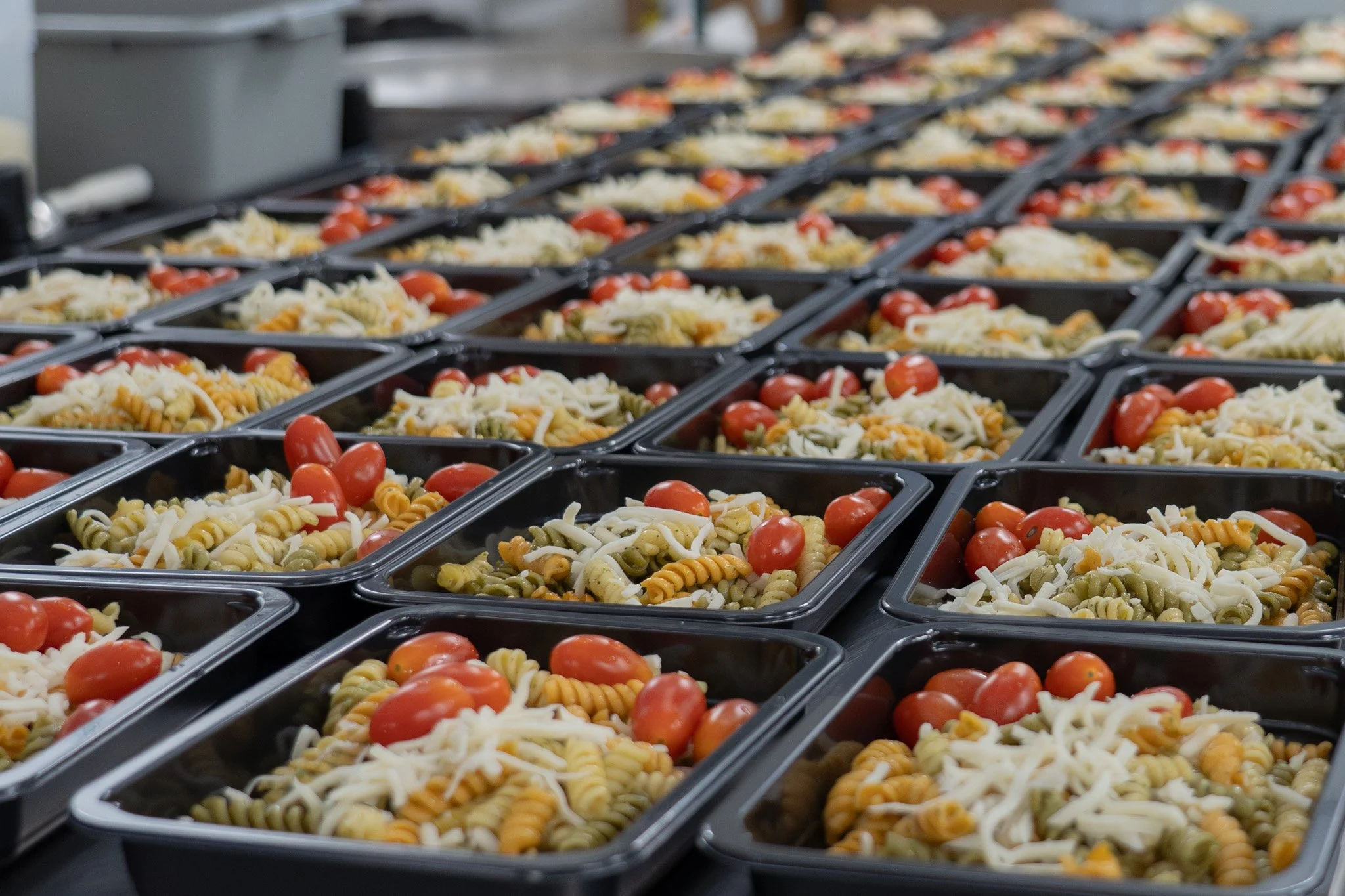 Food Consumption: Rows of black plastic meal trays containing rotini pasta with shredded cheese and grape tomatoes