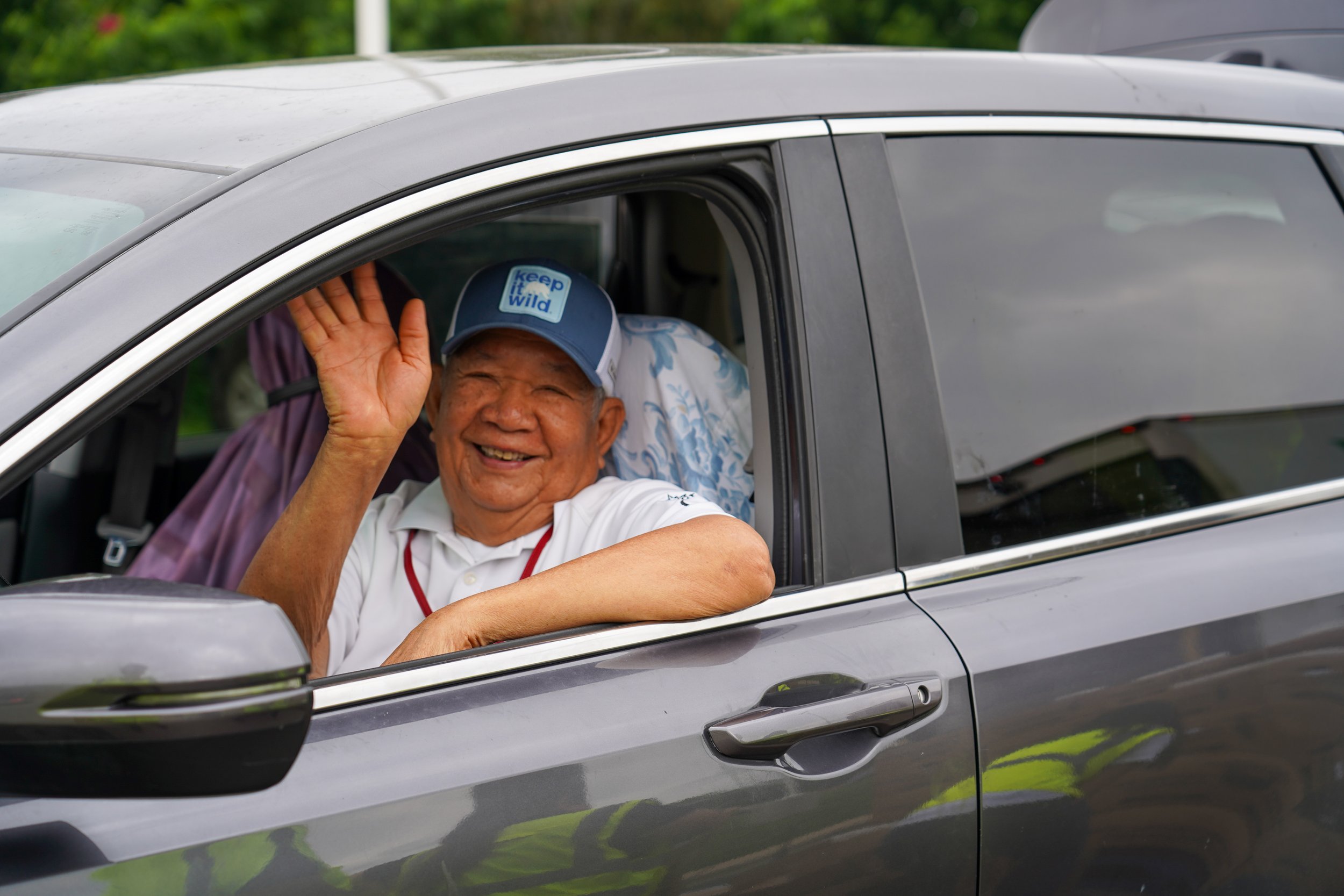 Smiling person in white shirt waving from driver's seat of gray vehicle