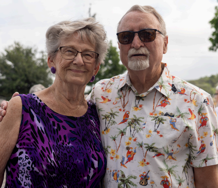 Person with sunglasses and multicolored bird shirt next to smiling person with glasses and purple top in front of tree