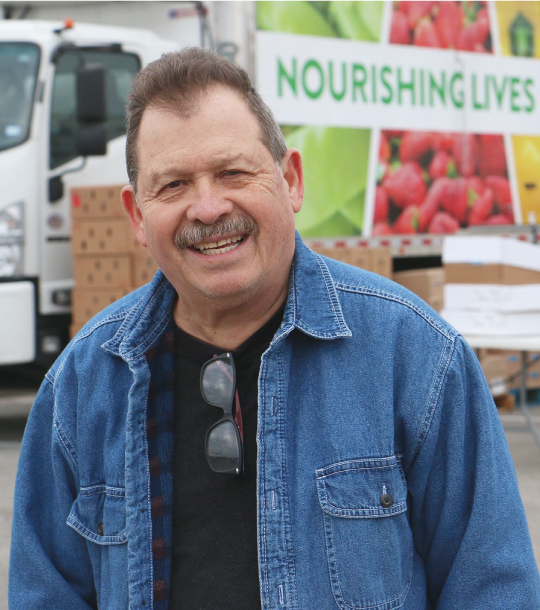 Smiling person in denim shirt standing in front of stacks of cardboard boxes and a Central Texas Food Bank truck