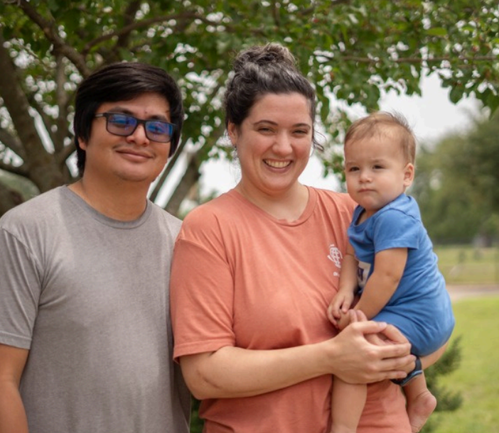 Smiling person in glasses and gray shirt next to smiling person in orange shirt holding baby in blue outside, with trees