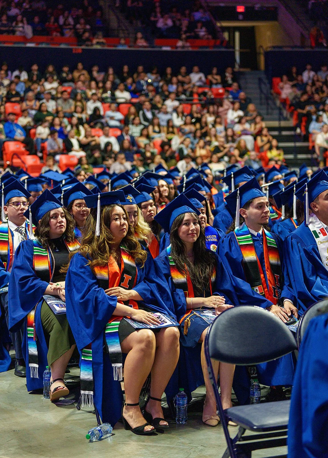 A large group of graduates wearing blue caps and gowns sit in rows at a graduation ceremony. Many graduates are wearing colorful stoles with patterns, and the audience is seated in the background.