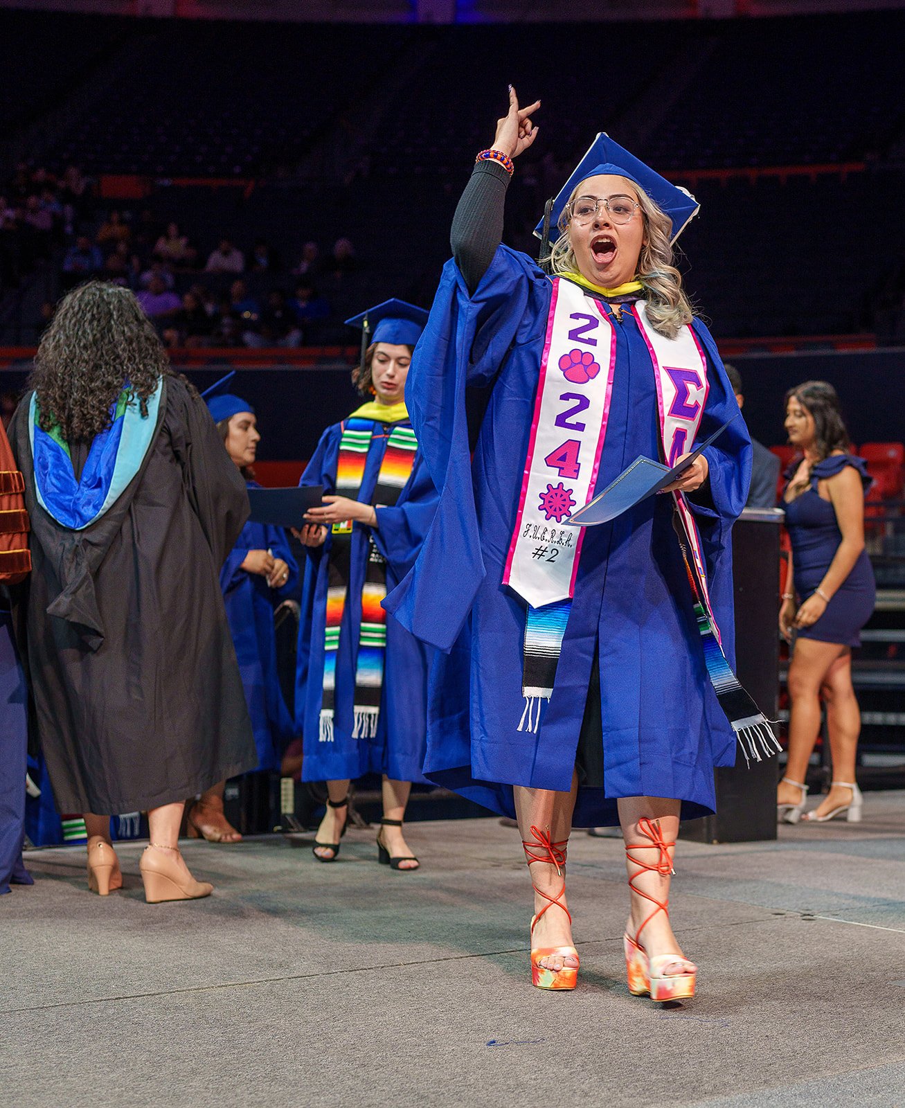 Group of graduates in blue caps and gowns, one celebrating, at a commencement ceremony with sashes displaying "2024".
