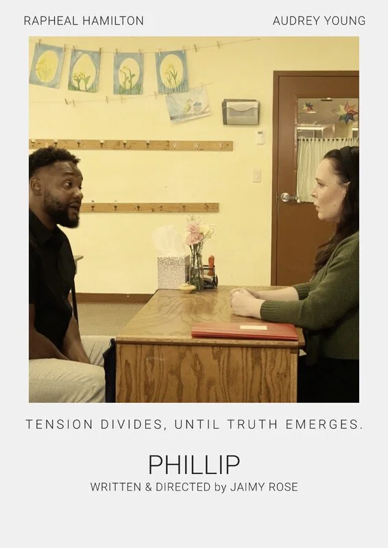 A man and a woman sit across from each other at a small wooden table in a room with yellow walls. The man has dark skin, short curly hair, and a beard. The woman has light skin and long, dark hair. There are flower decorations on the wall behind them, along with a string of artwork and a wooden coat rack. The table has a vase with pink flowers, a box of tissues, and a red folder.