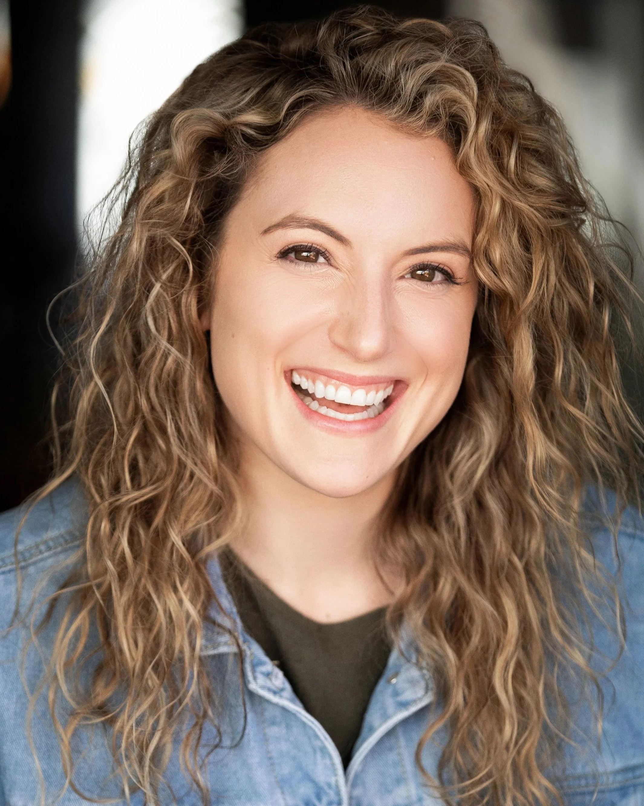 A smiling woman with curly hair wearing a denim jacket over a dark top.