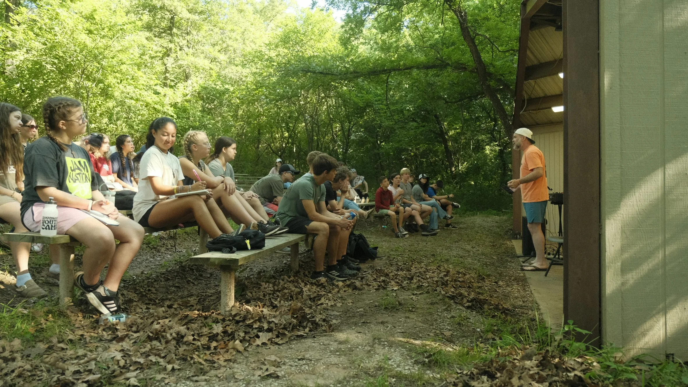 A group of young people sitting on benches outdoors, listening to a man speaking in front of a wooded area.