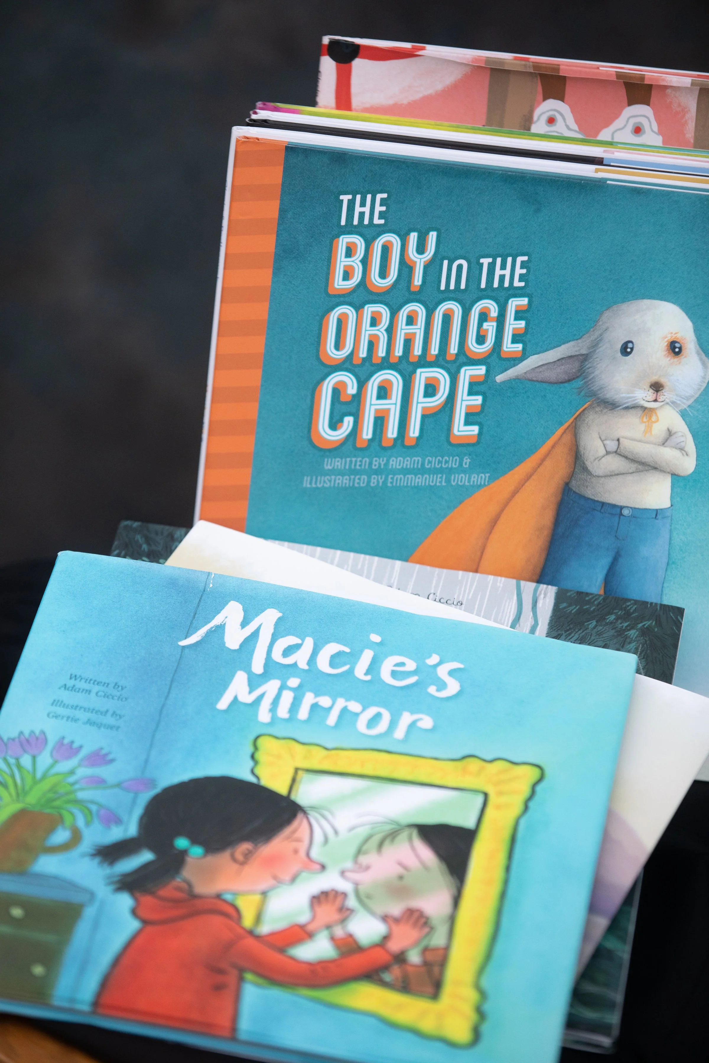 Several children's books stacked on a surface, with the top book titled 'The Boy in the Orange Cape' featuring an illustrated character of a boy with a rabbit-like face, crossed arms, and an orange cape.