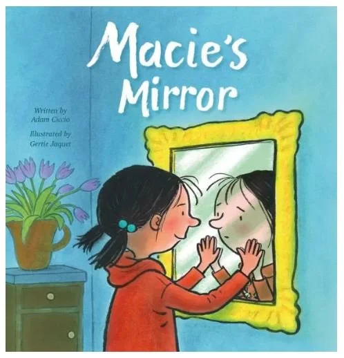 Cover of a children's book titled 'Macie's Mirror' showing a girl with dark hair and a red sweater looking at her reflection in a mirror and touching it, with a vase of purple flowers on a green table in the background.