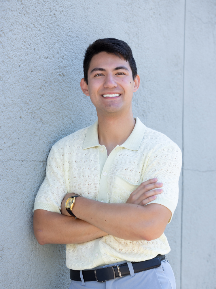 A young man with short dark hair smiling and standing with crossed arms against a light gray textured wall, wearing a cream short-sleeve polo shirt, gray pants, and a gold watch.