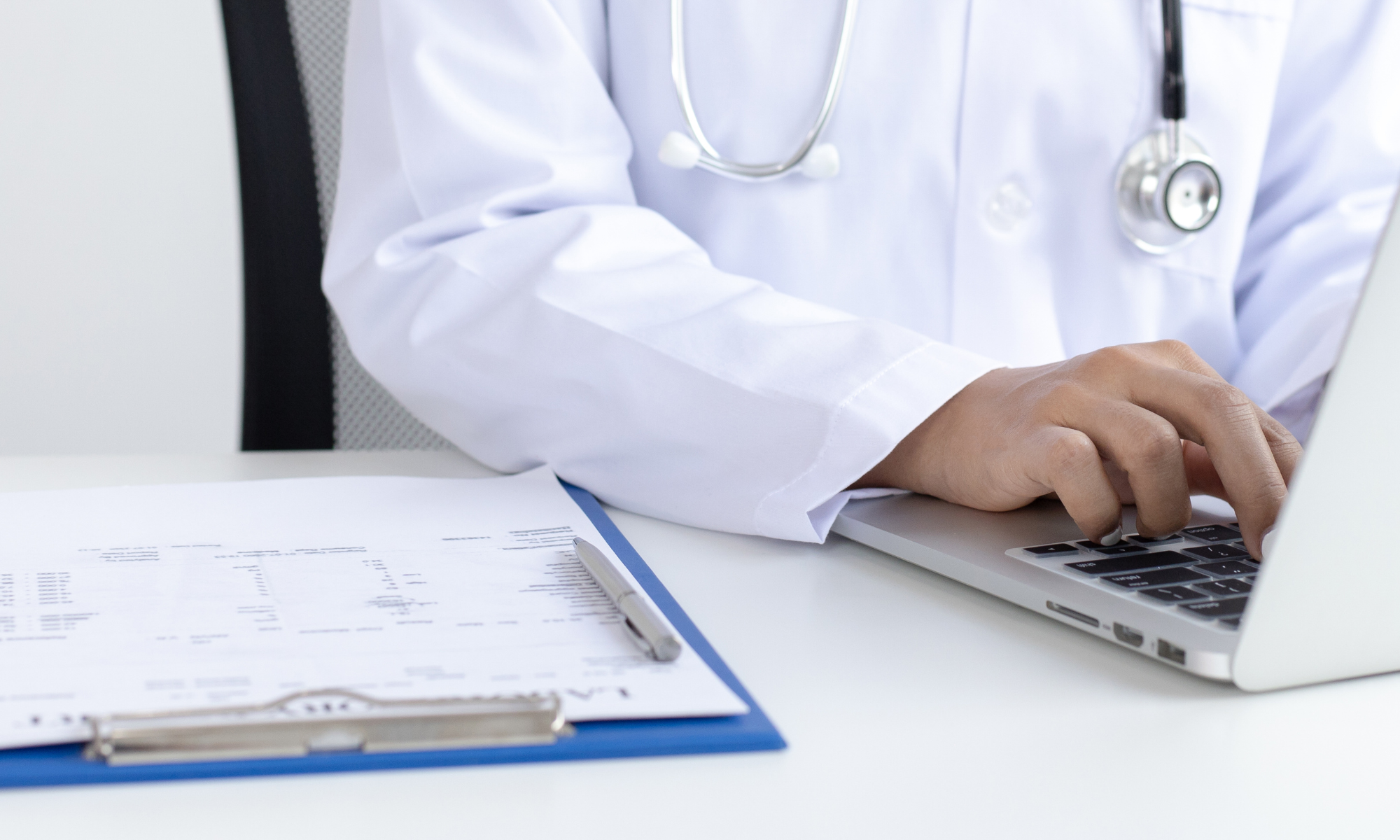 A person wearing a white medical coat and a stethoscope on their neck working on a laptop at a desk with documents and a pen.