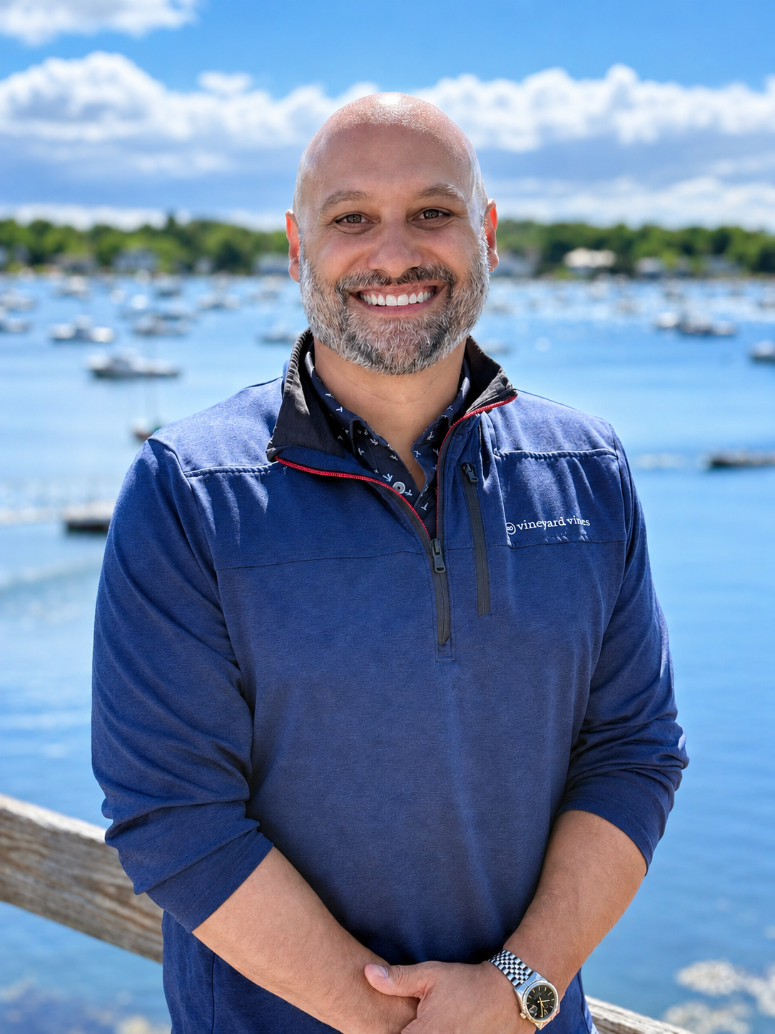 Smiling man with a beard and bald head standing outdoors near a body of water with boats, wearing a blue jacket and a watch, under a partly cloudy sky.