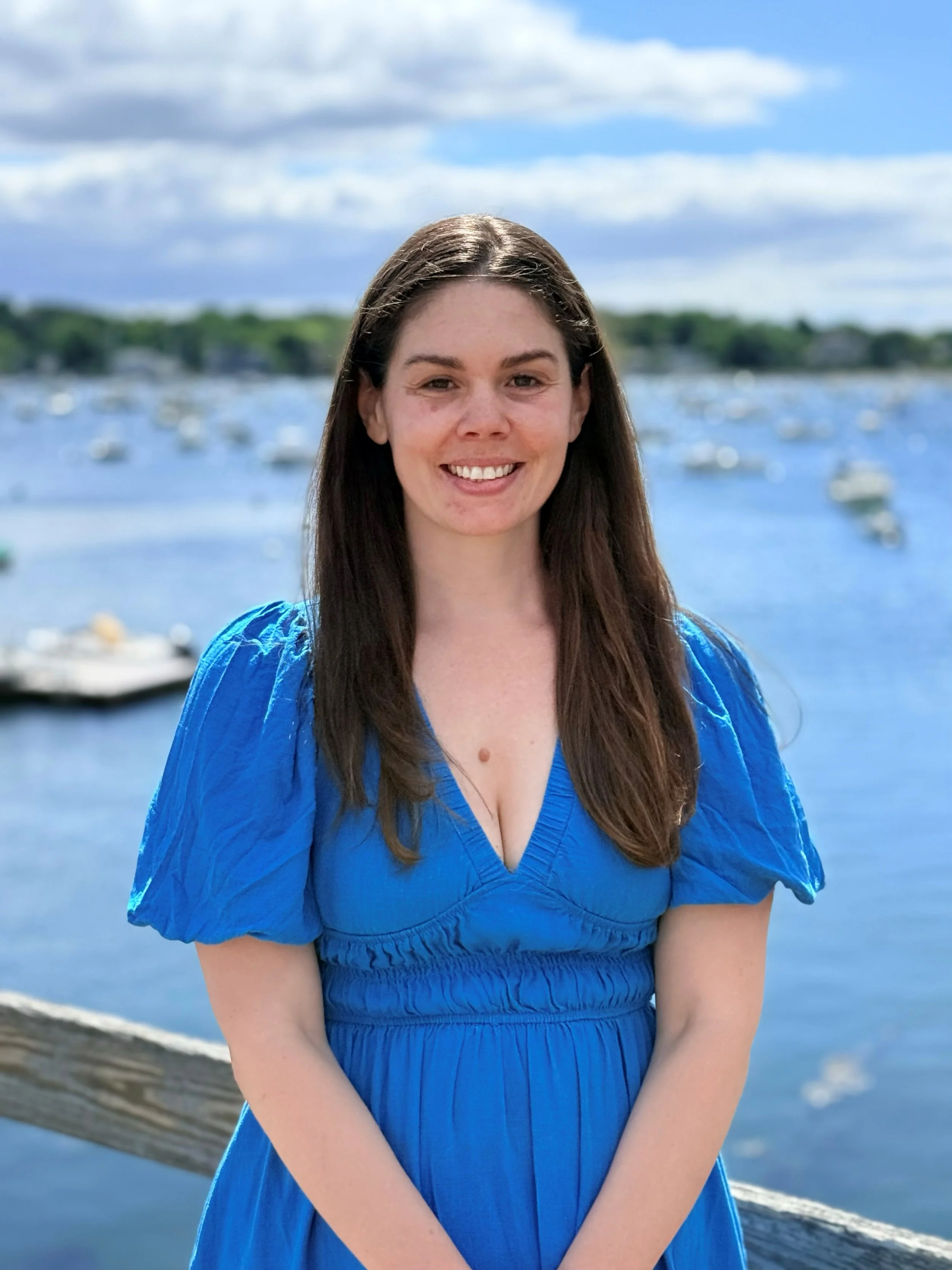 A woman with long brown hair smiling outdoors near a body of water, wearing a blue dress, with a cloudy sky and green trees in the background.