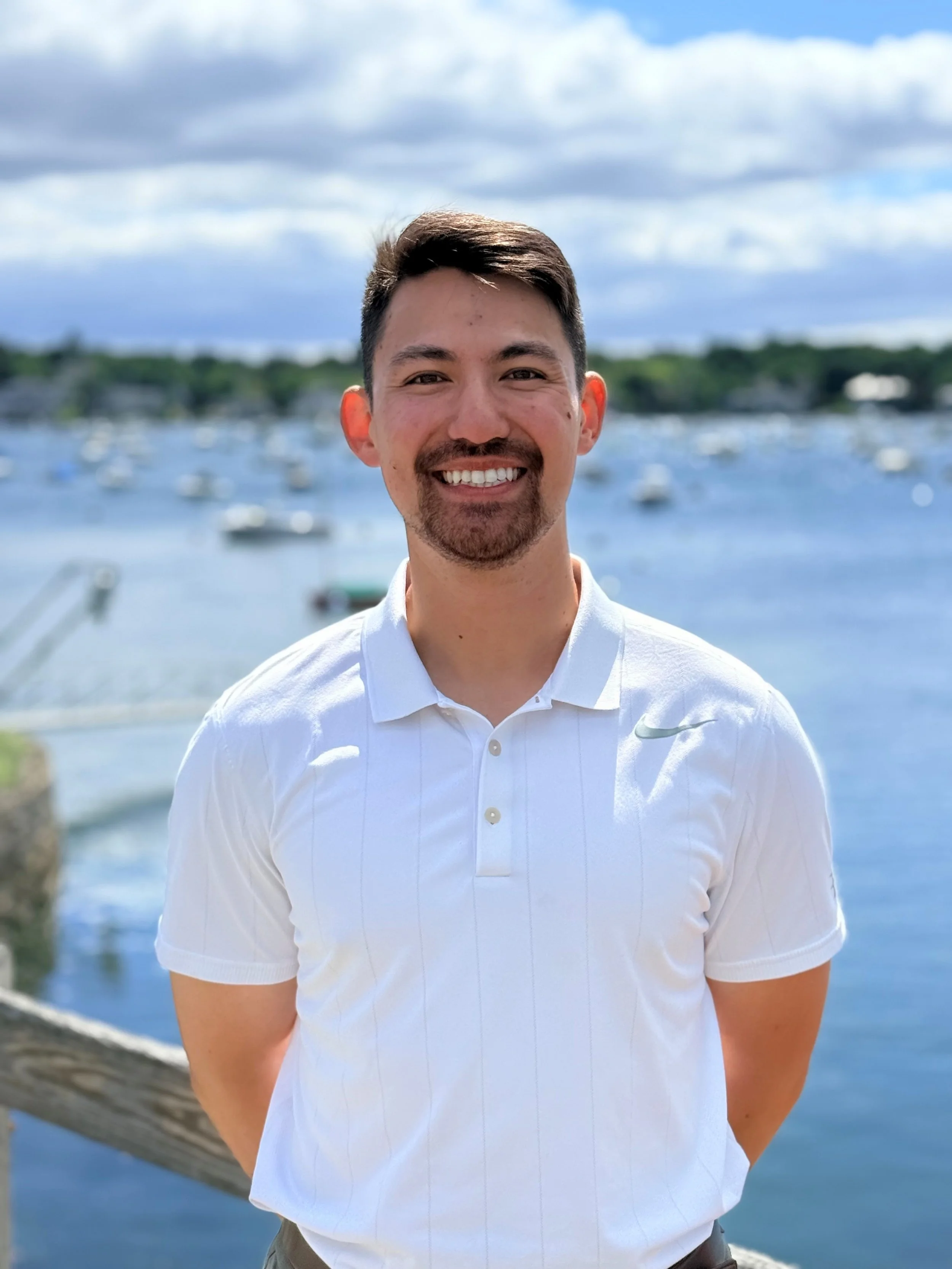 A smiling man with short dark hair and a goatee stands outdoors near a body of water with boats and a tree-lined shoreline in the background on a partly cloudy day.
