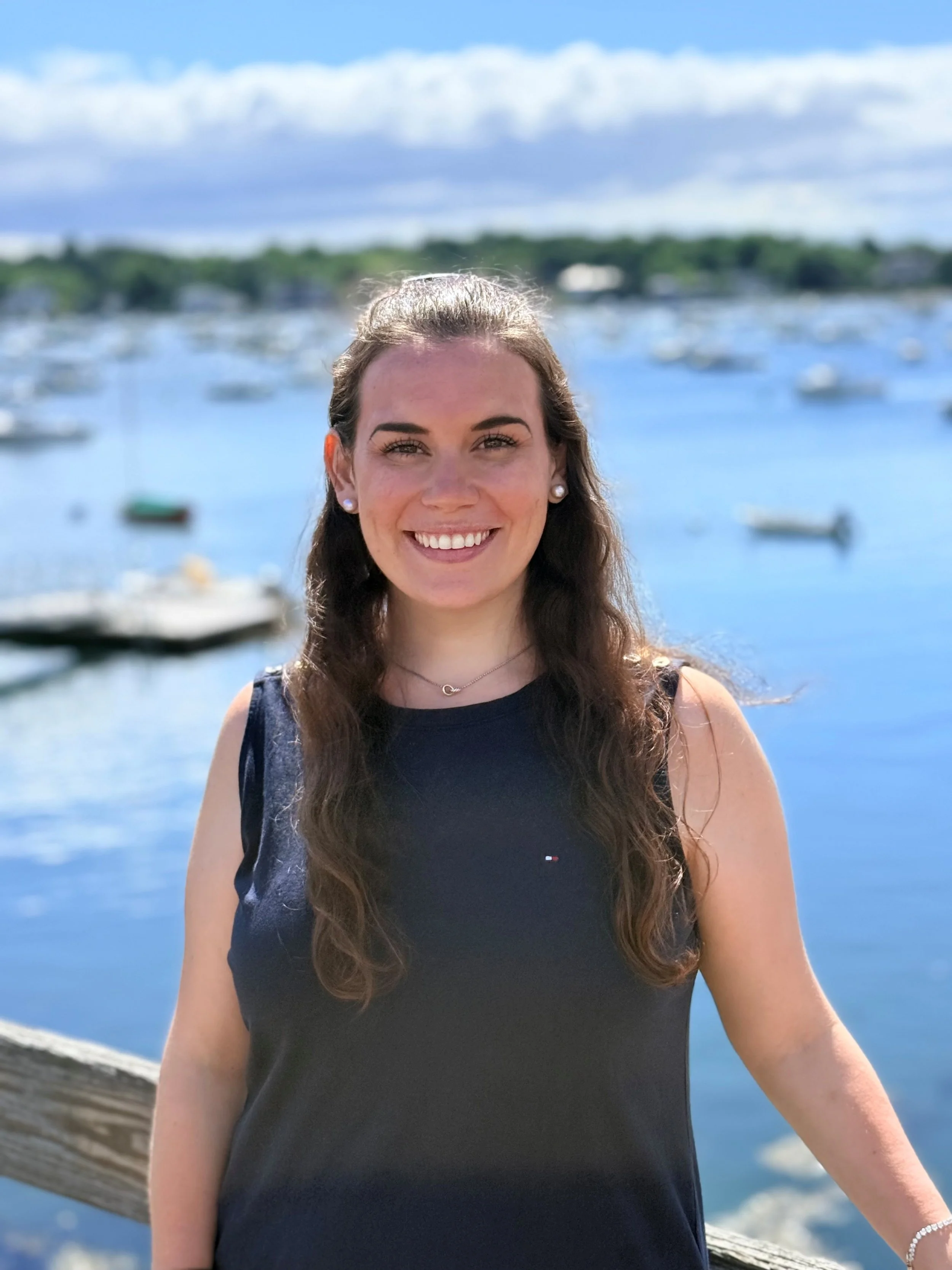 A smiling woman with long brown hair wearing a black sleeveless top, earrings, and a necklace, standing outdoors near a body of water with boats and greenery in the background on a sunny day.