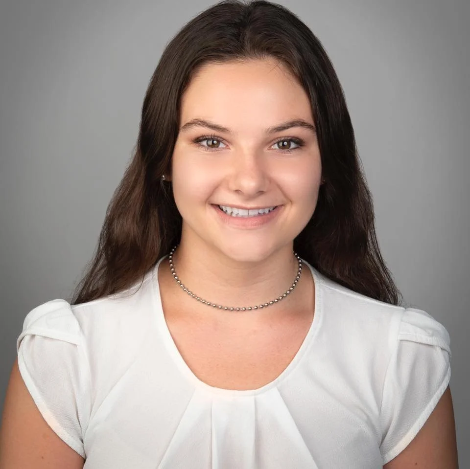 A young woman with long brown hair, wearing a white top and a silver beaded necklace, smiling against a gray background.