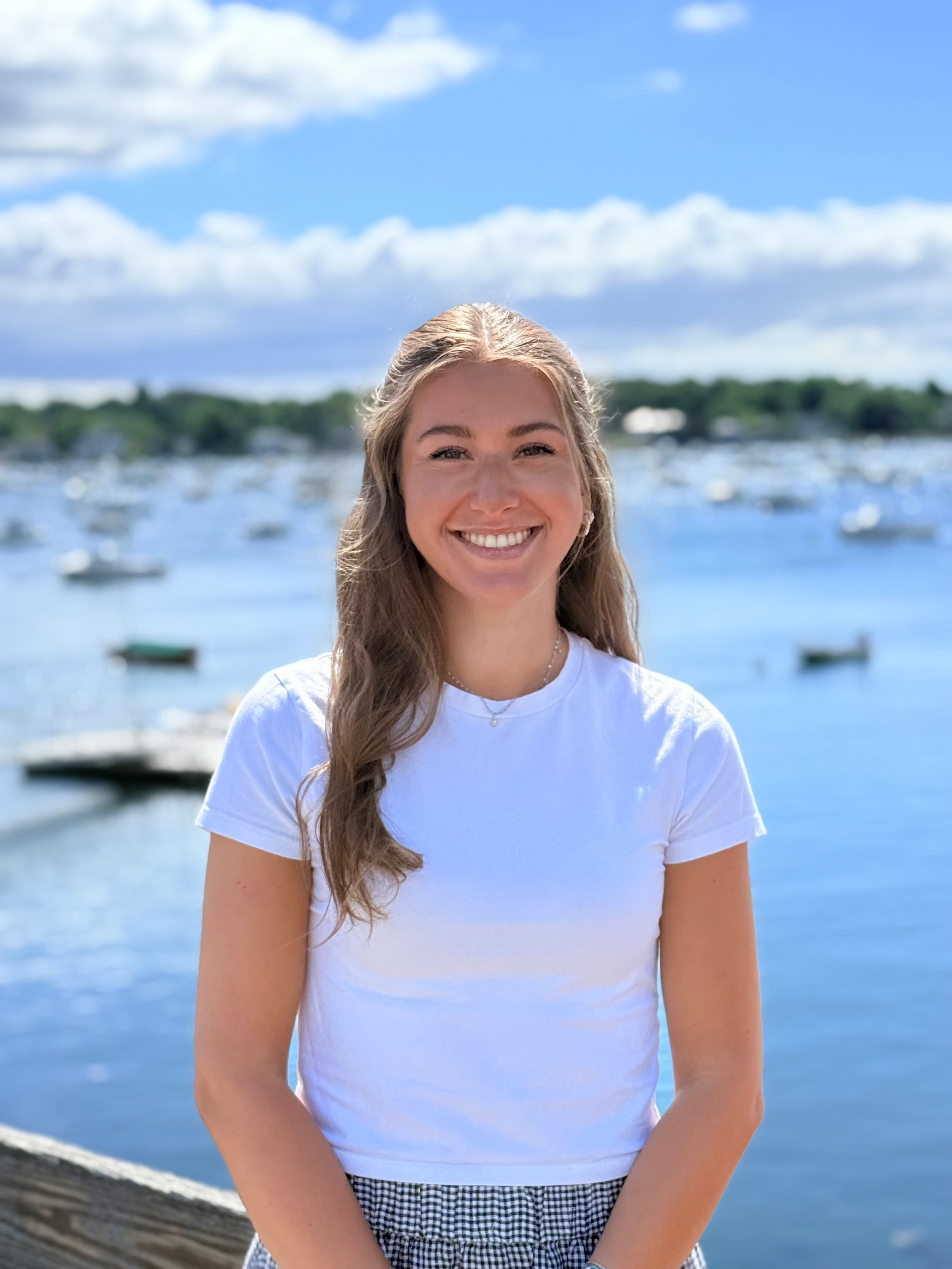 Young woman smiling at the camera by a body of water with boats and a tree-lined shoreline in the background on a sunny day.