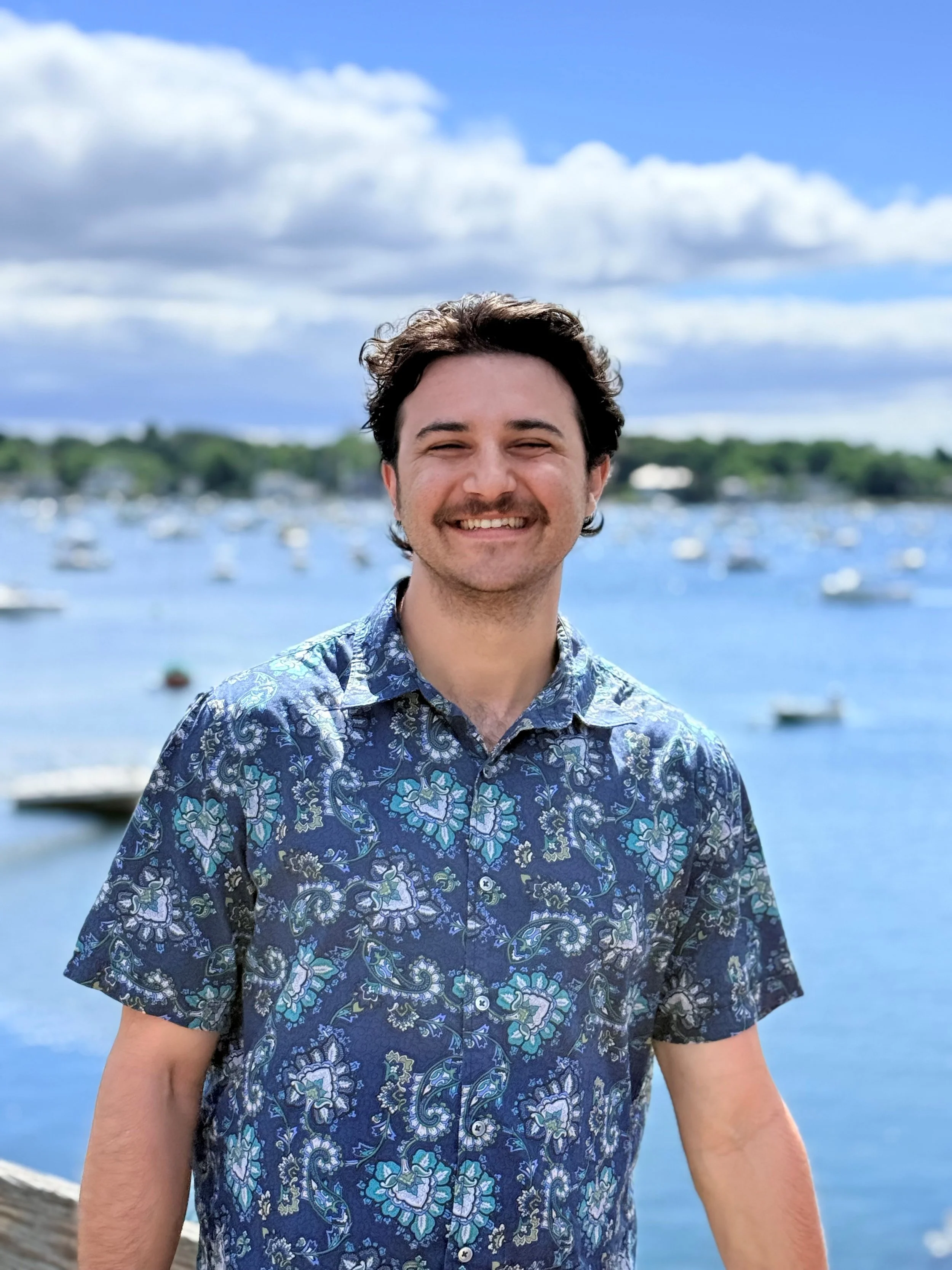 A smiling man in a patterned shirt standing near a body of water with boats and greenery in the background on a partly cloudy day.