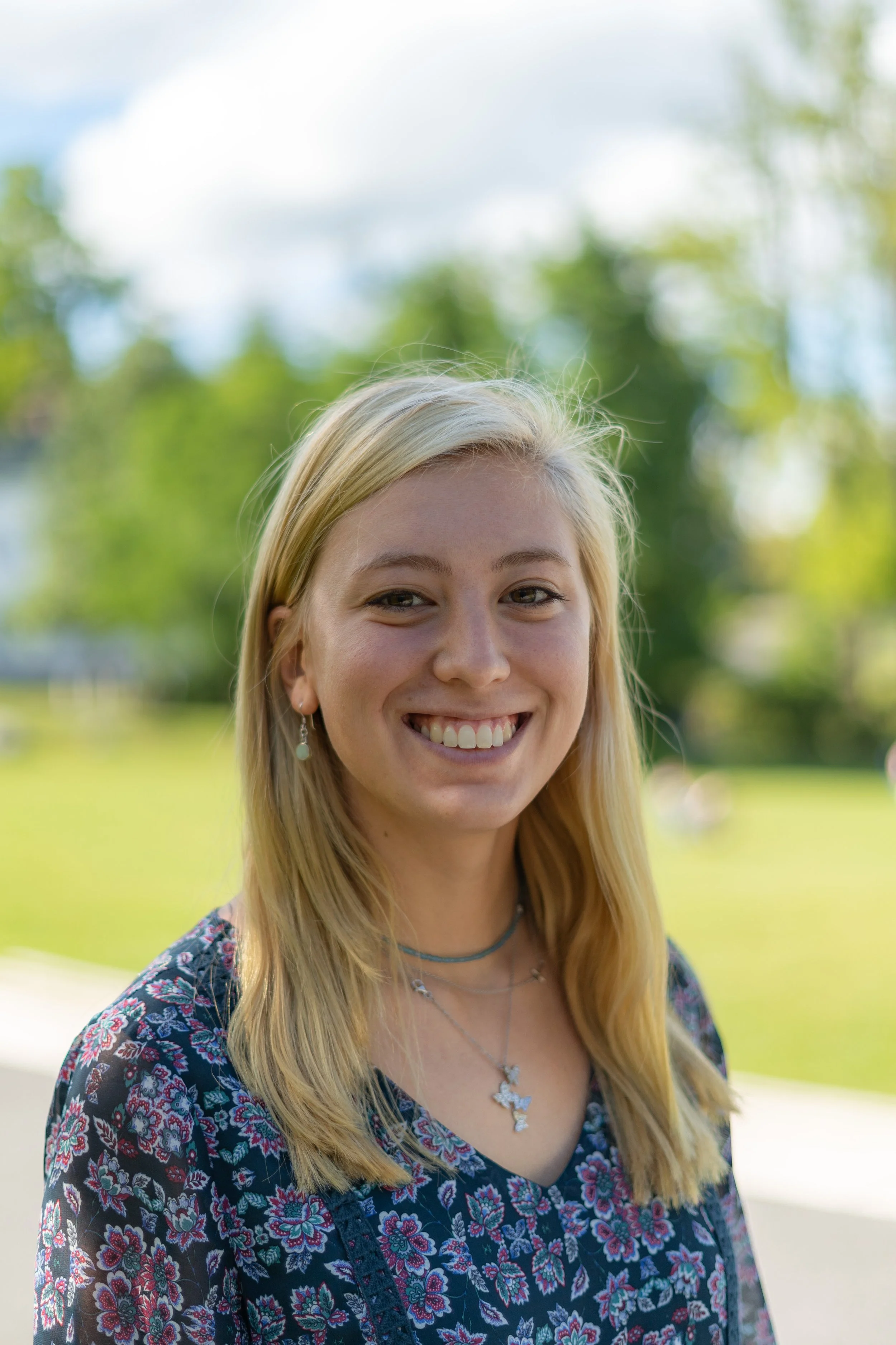 Young woman with blonde hair smiling outdoors in a park with green trees and blue sky in the background.