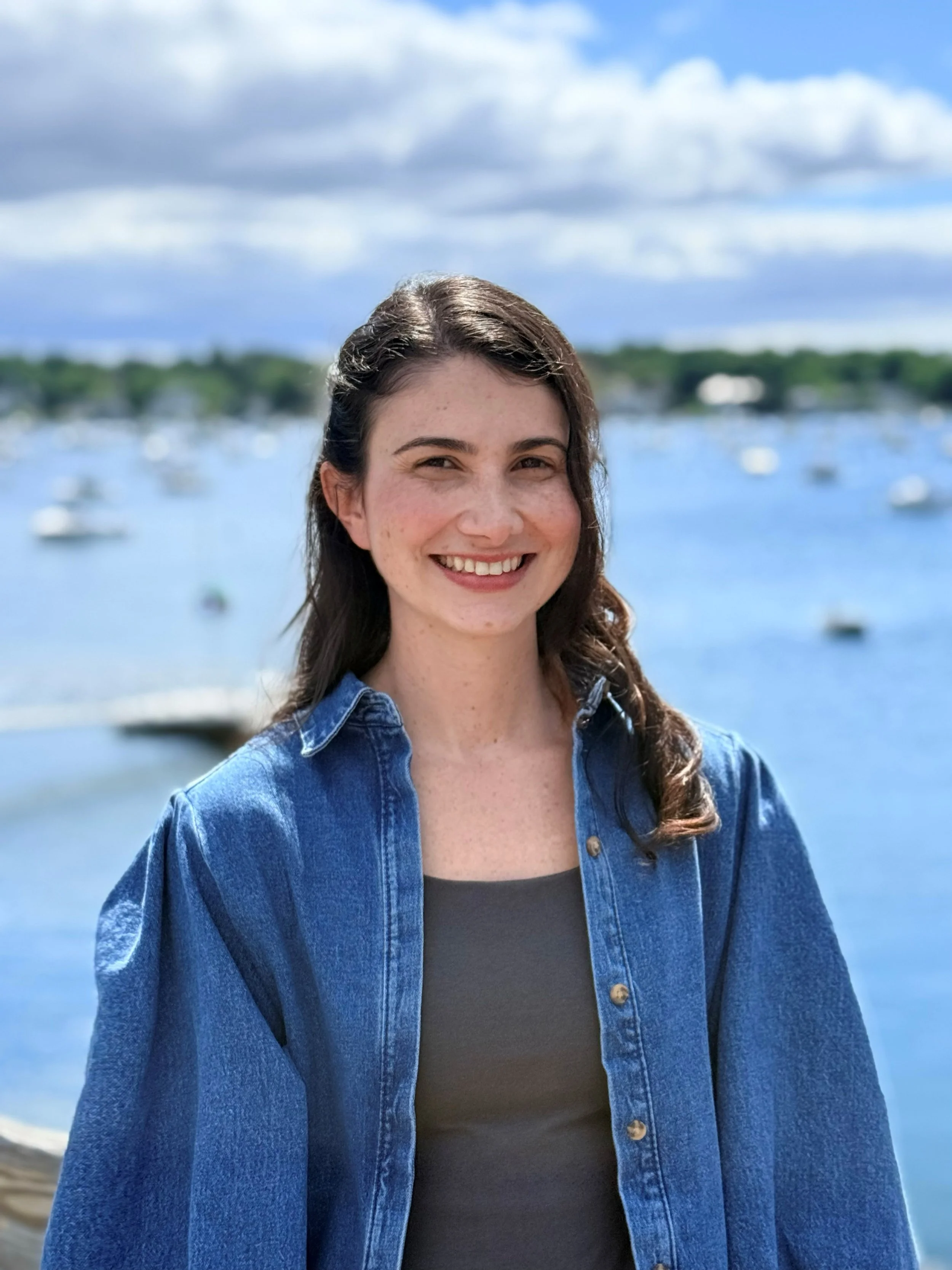 A young woman with long, wavy brown hair smiling at the camera, wearing a blue denim jacket over a black top, with a body of water and boats in the background under a partly cloudy sky.