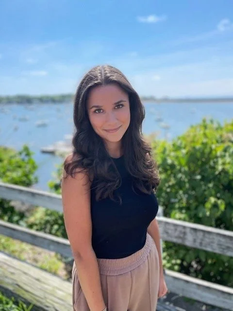 Young woman with long dark hair standing outdoors near a water body, with a blue sky and greenery in the background.