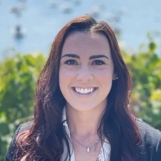 A woman smiling outdoors with greenery and water in the background.