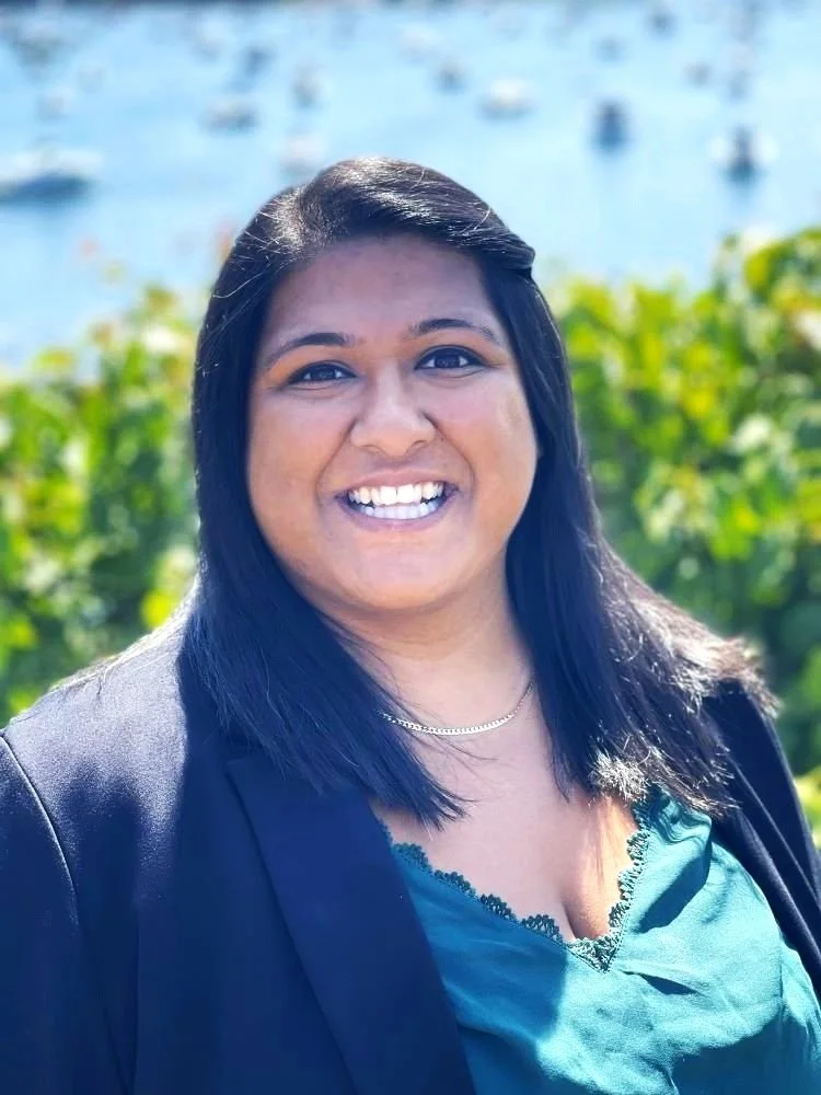 A woman smiling outdoors with a blurred background of water and boats, wearing a dark blazer and teal top.