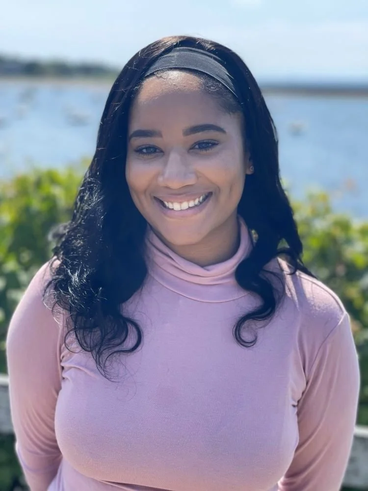 Smiling woman with long dark hair, black headband, and pink turtleneck sweater outdoors near a body of water and green bushes.