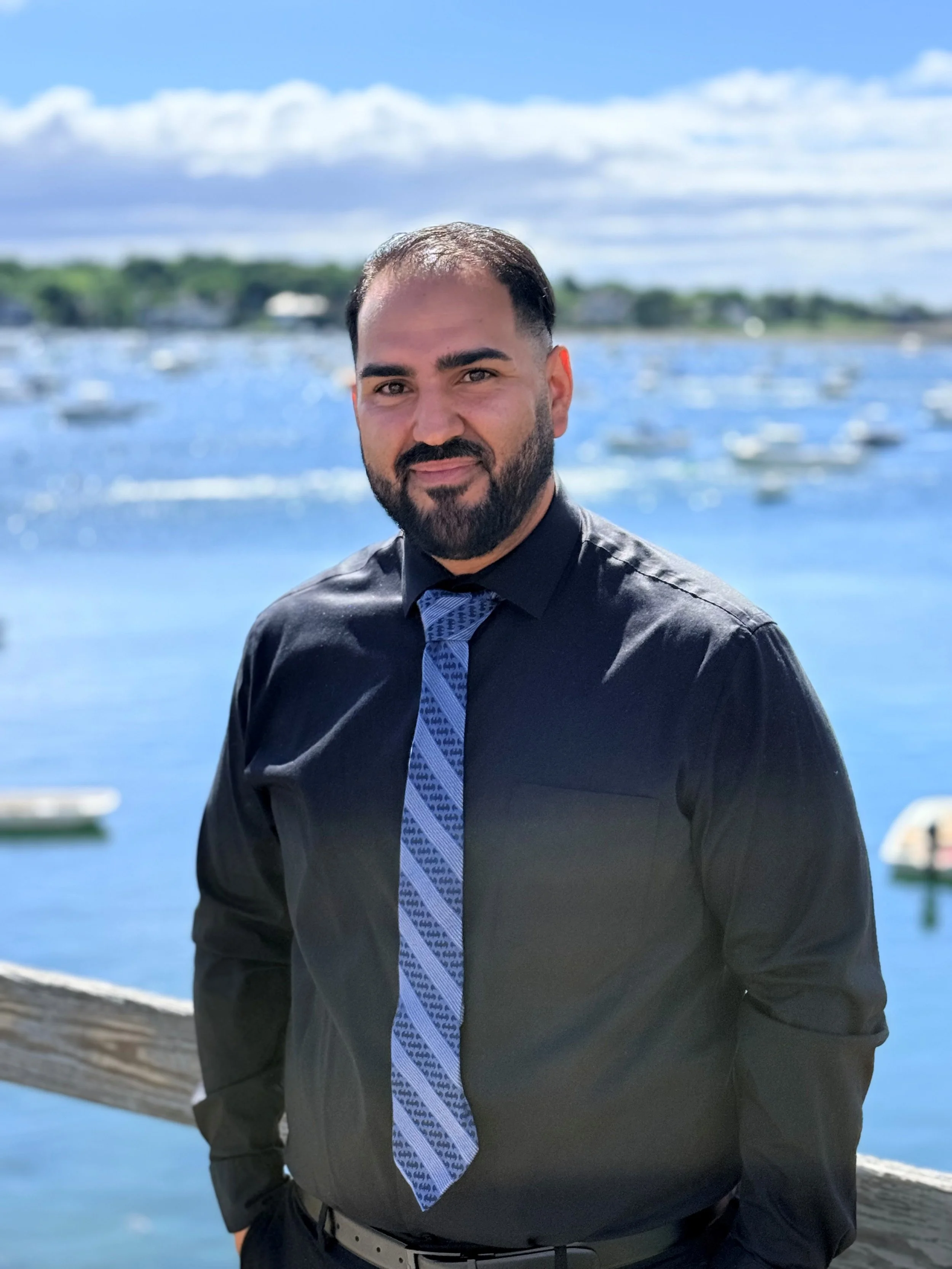 A man with a beard and short dark hair standing outdoors near a body of water with boats, under a partly cloudy blue sky, wearing a black shirt and a patterned tie.