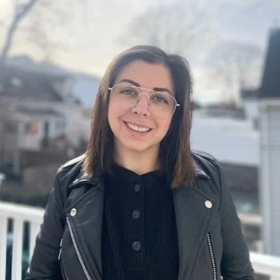 Young woman with shoulder-length brown hair, glasses, smiling, wearing a black jacket, standing outdoors in a residential neighborhood.