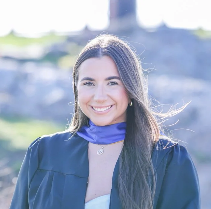 Young woman in graduation gown and blue stole smiling outdoors with blurred background.