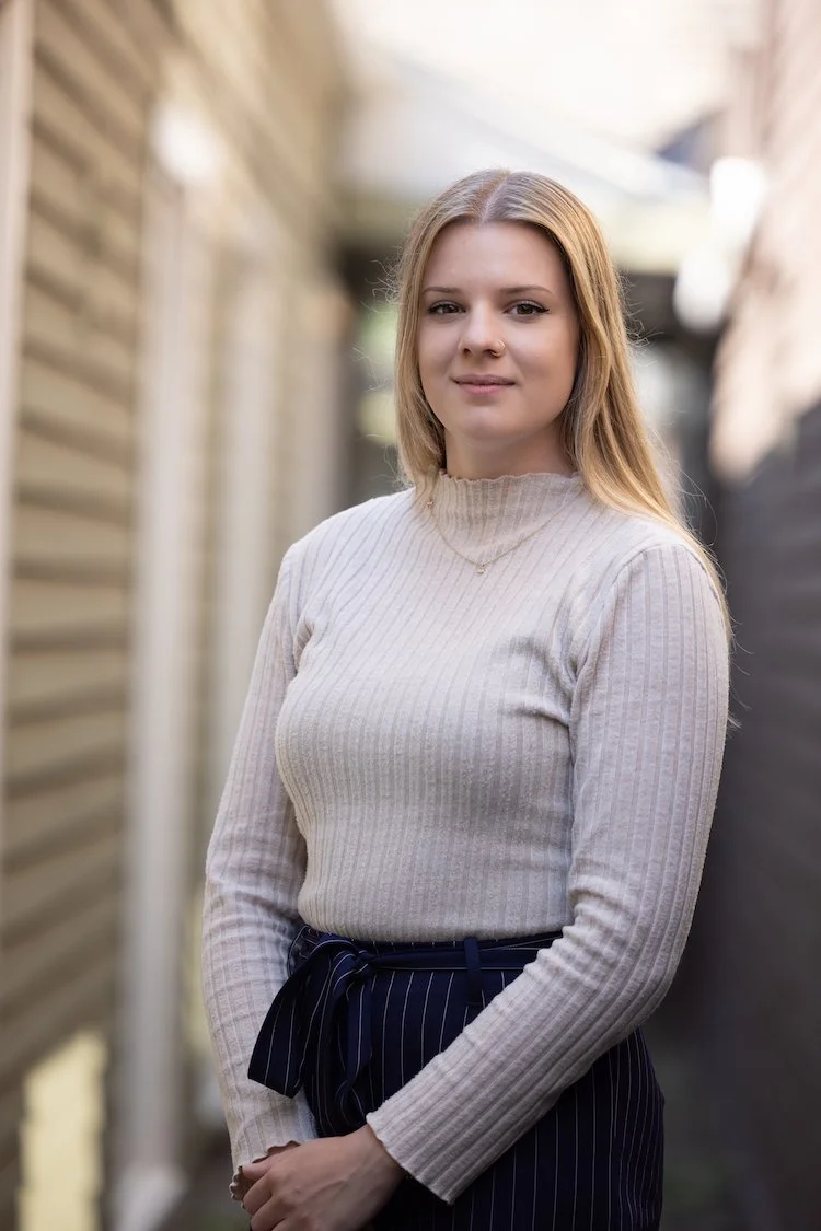A young woman with light brown hair, wearing a beige ribbed turtleneck sweater and navy pinstripe pants, standing outdoors with a blurred background of a wooden fence and buildings.