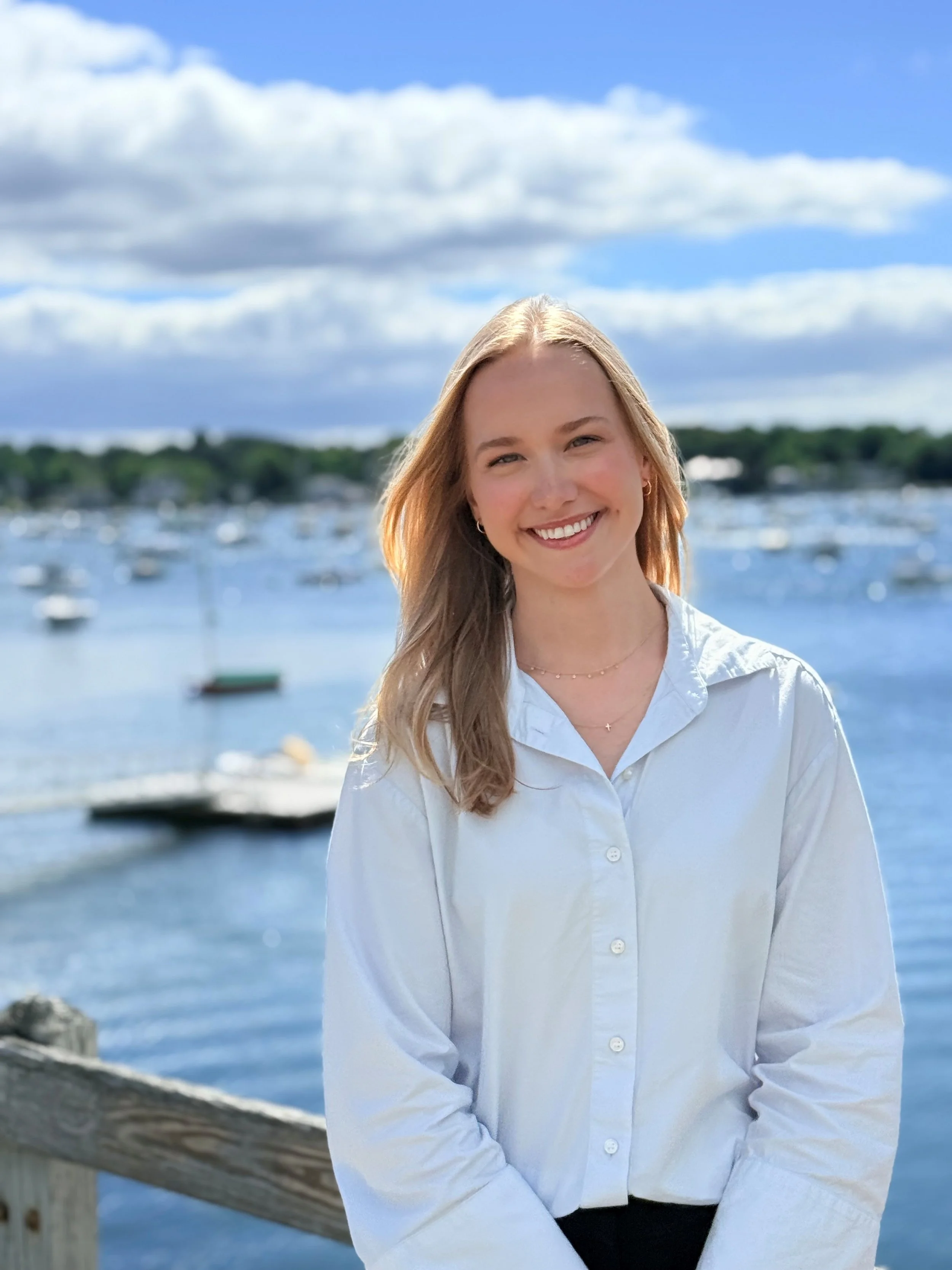 A young woman with blonde hair and a white button-up shirt smiling in front of a harbor with boats and a blue sky with clouds.