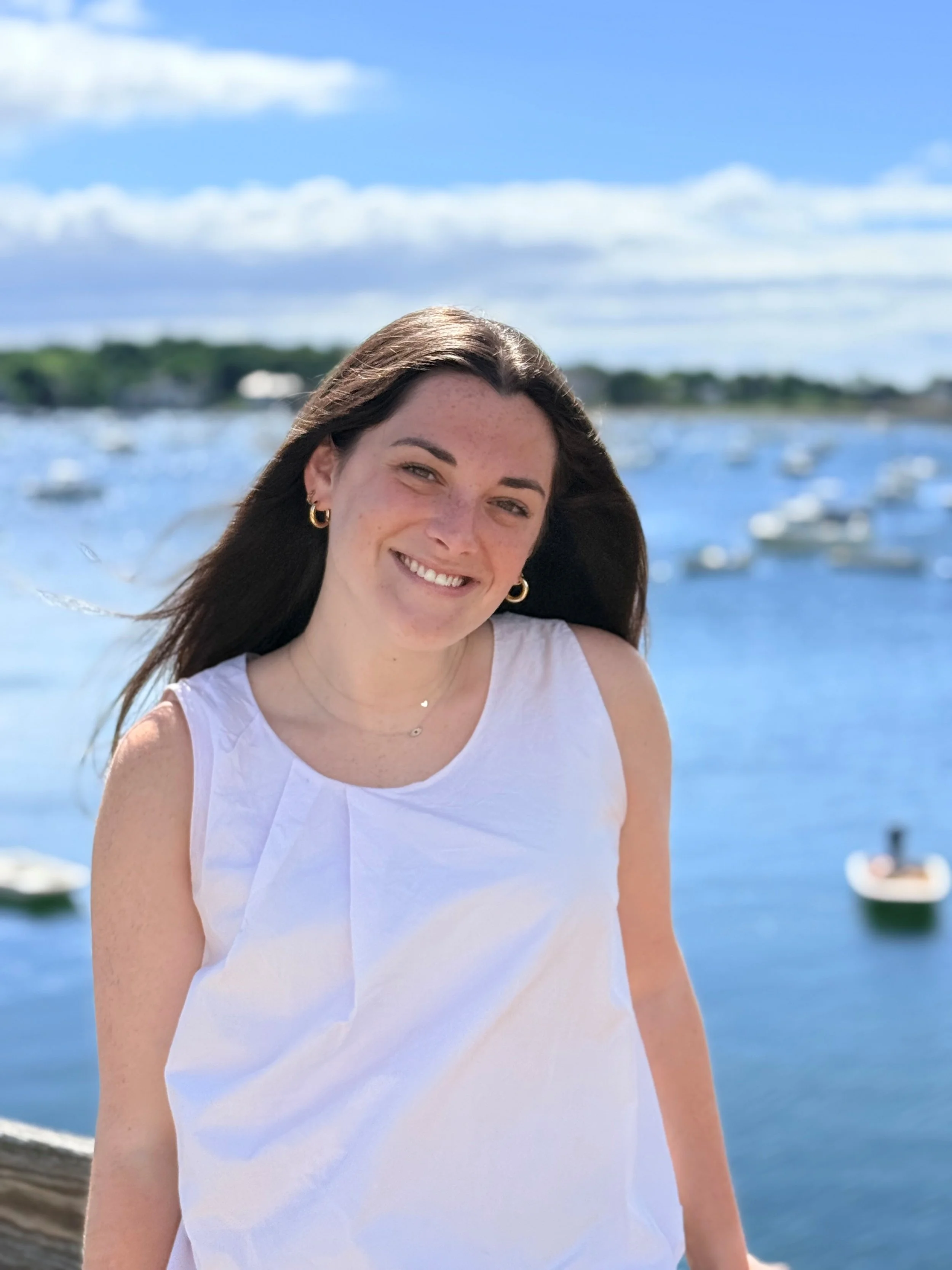 Smiling woman with long brown hair, wearing earrings and a white sleeveless top, standing outdoors near water with boats and a shoreline in the background.