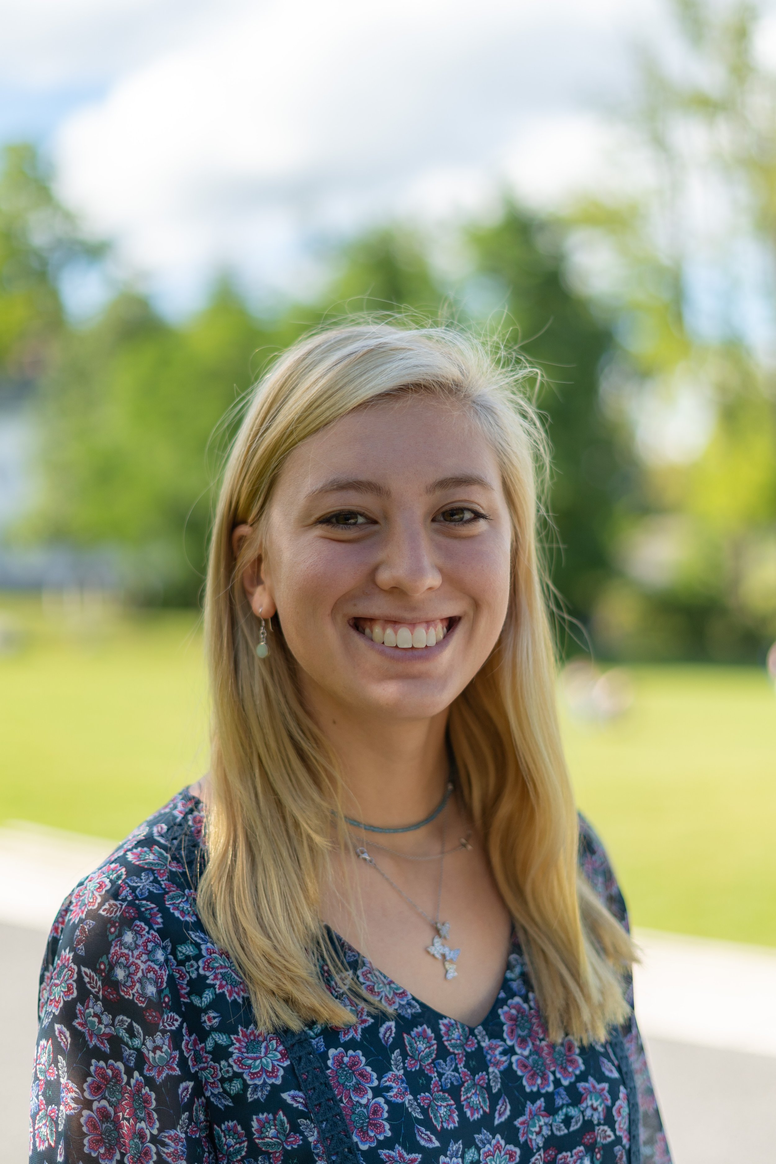Young woman with blonde hair smiling outdoors, wearing a floral-patterned dress and jewelry, with green trees and a bright sky in the background.