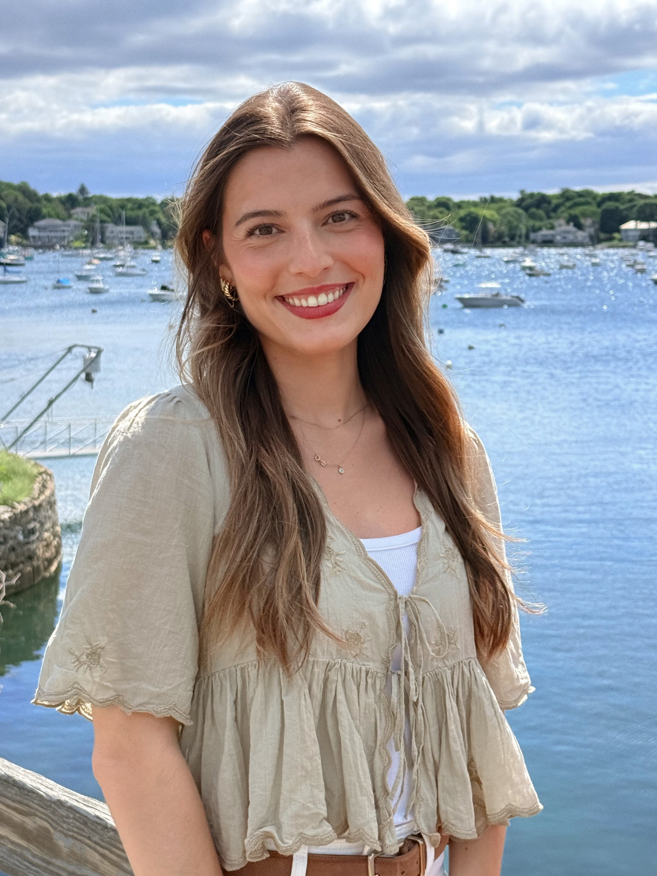 Smiling woman with long brown hair standing outdoors near a body of water with boats and houses in the background on a partly cloudy day.