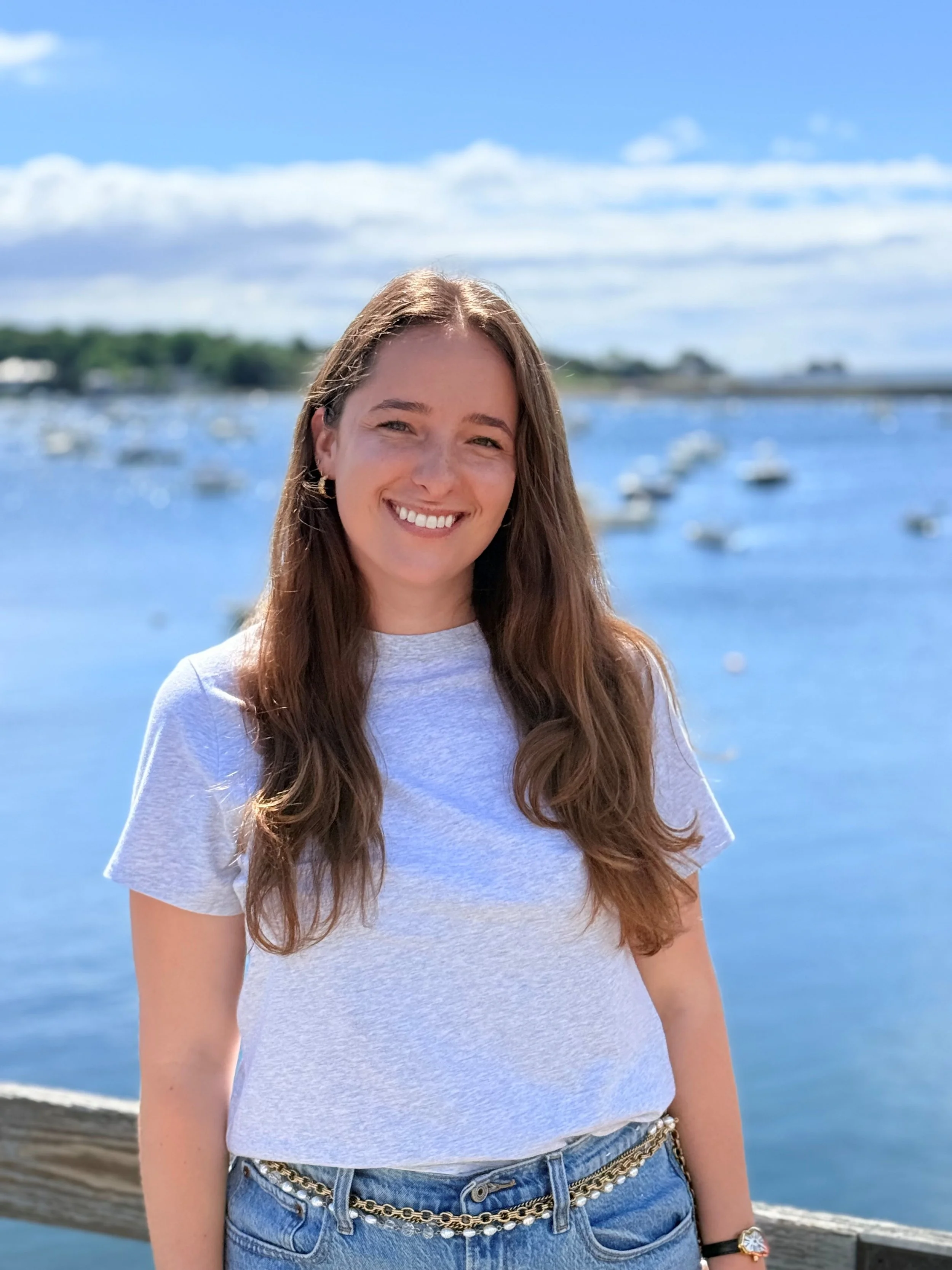 Young woman with long brown hair smiling outdoors near a body of water with boats and a blue sky in the background.