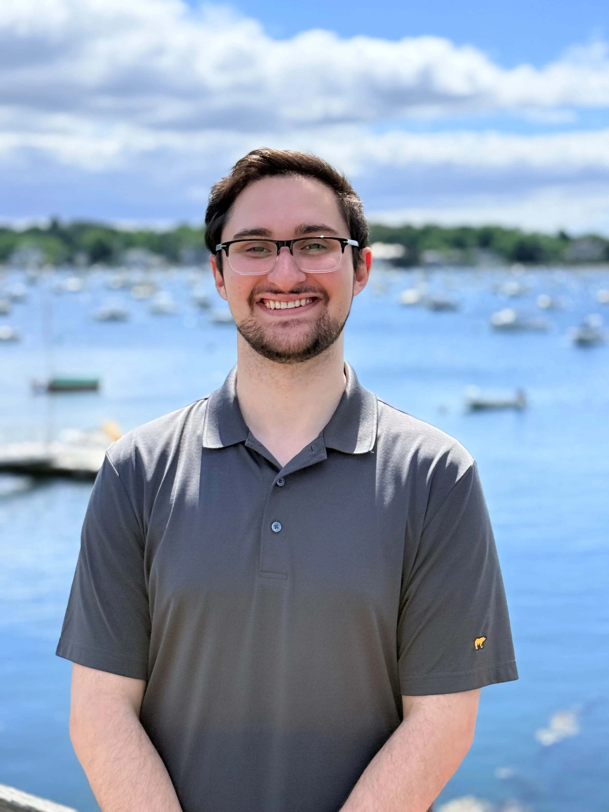A smiling young man with glasses, wearing a gray polo shirt, standing outdoors near a body of water with boats, trees, and a cloudy sky in the background.
