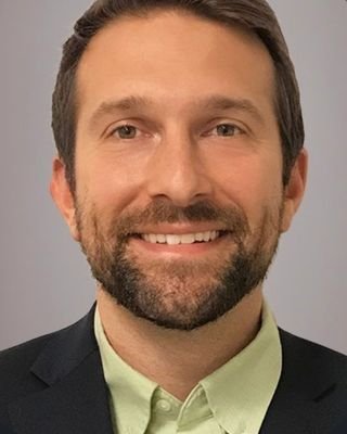 A smiling man with dark hair and a beard, wearing a dark suit jacket and a light green dress shirt, against a neutral background.