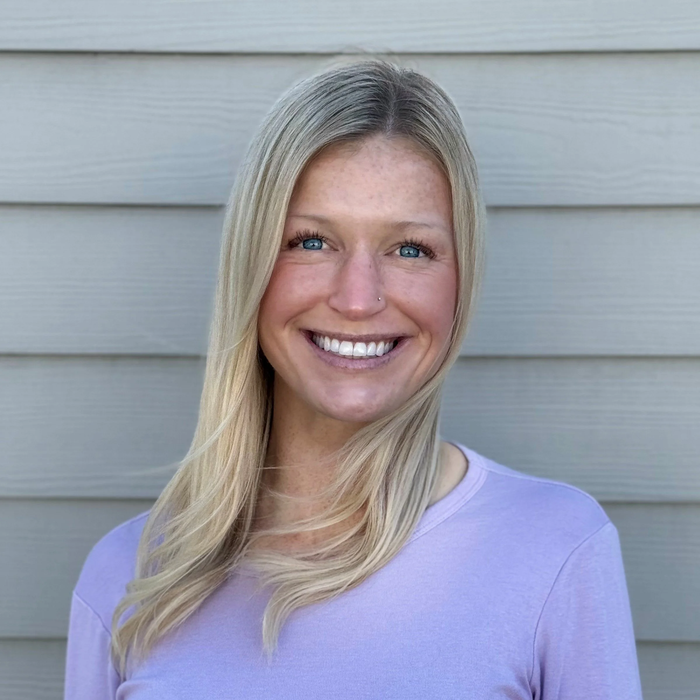 Close-up of a woman with long blonde hair, blue eyes, and a small nose piercing, wearing a pastel purple top, smiling in front of gray horizontal siding.