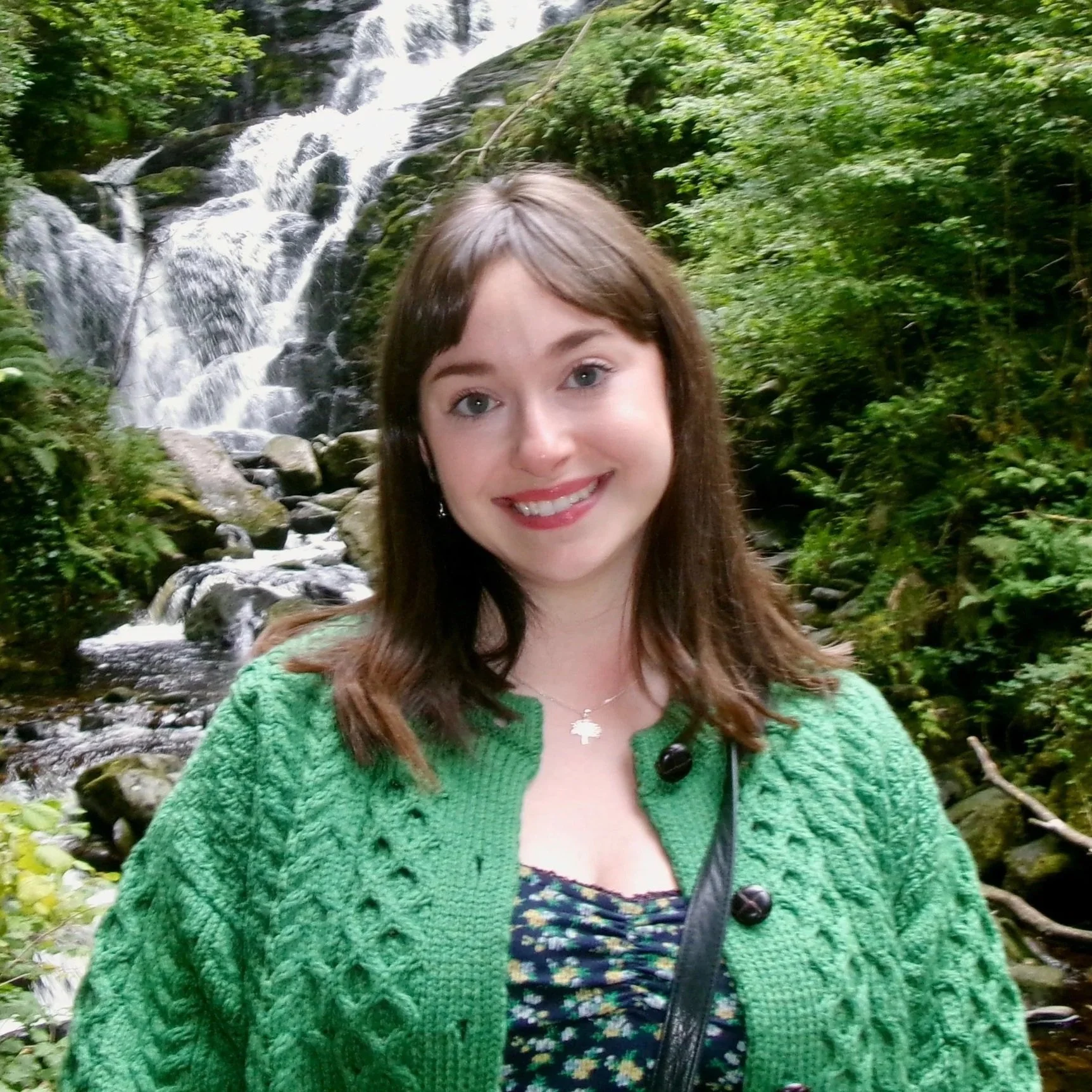 A woman with brown hair and blue eyes smiling in front of a waterfall surrounded by green foliage.