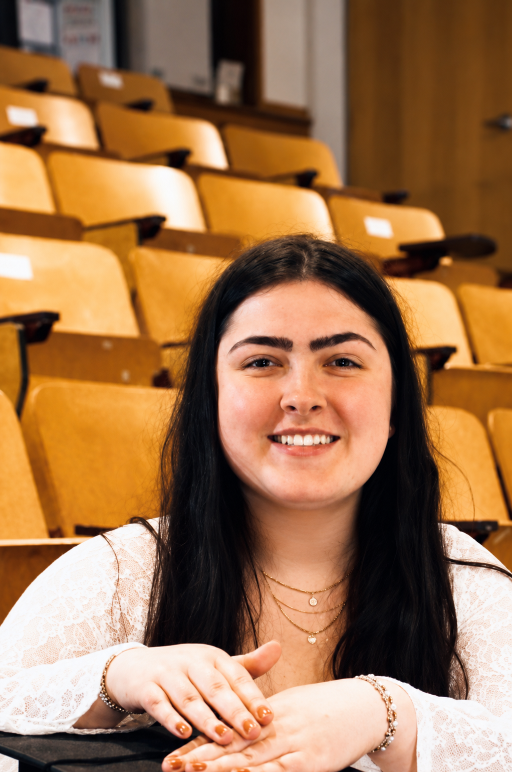 A woman with long black hair sitting in an auditorium with empty yellow seats, smiling at the camera.